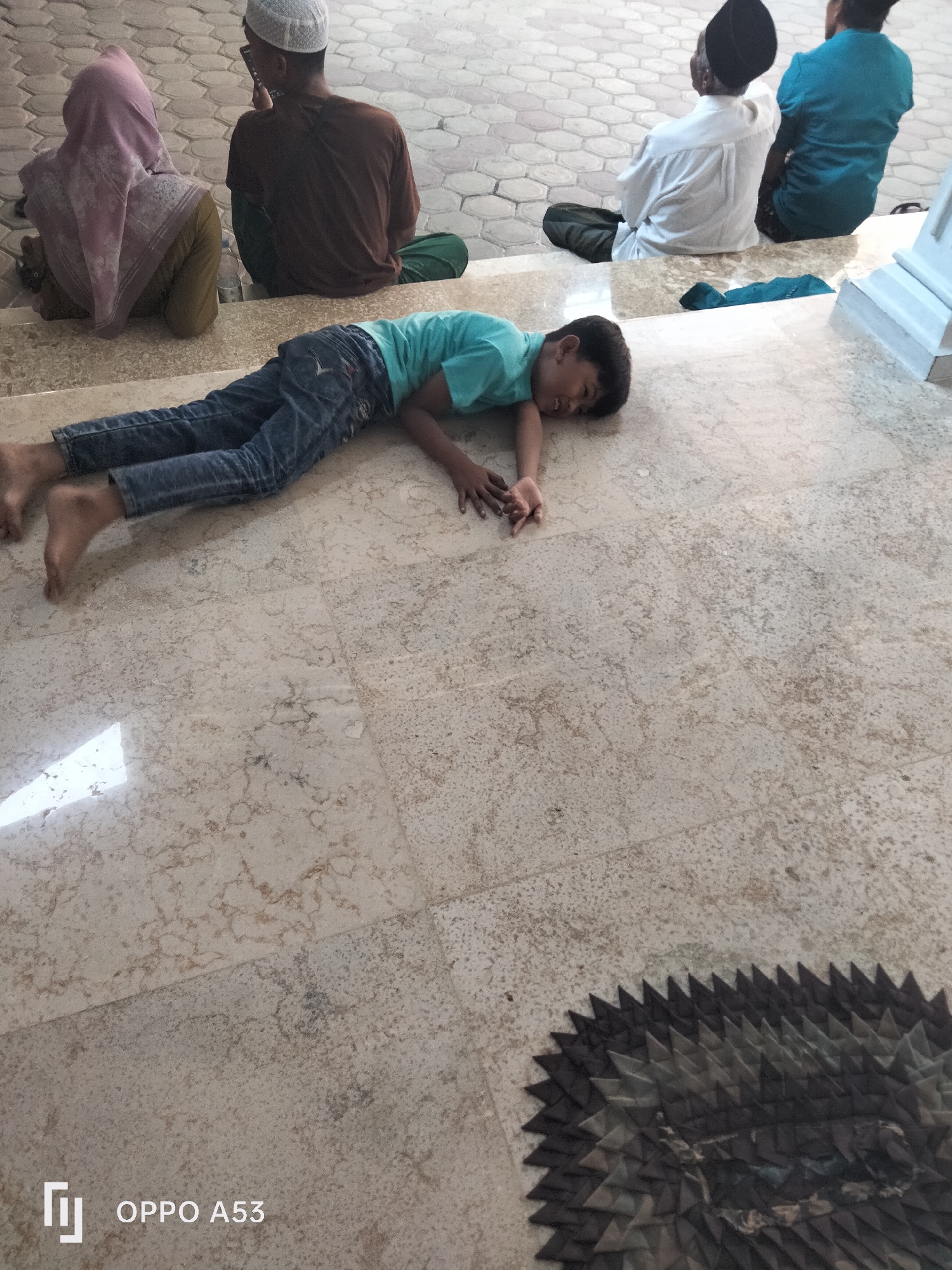 In an outdoor setting, possibly a public space in Badung, Indonesia, a young boy is lying on his stomach on a marble-tiled floor, appearing to be asleep or resting. He is wearing a teal t-shirt and blue jeans, with his bare feet visible. His body is positioned horizontally across the frame, with his head turned to his right, resting on his arm.
In the background, several individuals are seated on steps and the floor, facing away from the camera. One person wears a pink hijab and a green garment, another a brown t-shirt and a white skullcap, and two others are in white and blue attire, respectively, with one wearing a black cap. One of the seated individuals is looking at a phone. A dark, decorative mat with a geometric pattern is visible in the lower right corner of the image. The flooring appears to be polished marble tiles with a light beige and brown veining pattern. The lighting suggests it is daytime. A white structure, possibly a pillar or part of a building, is visible on the far right. The "OPPO A53" watermark is present in the bottom left corner.