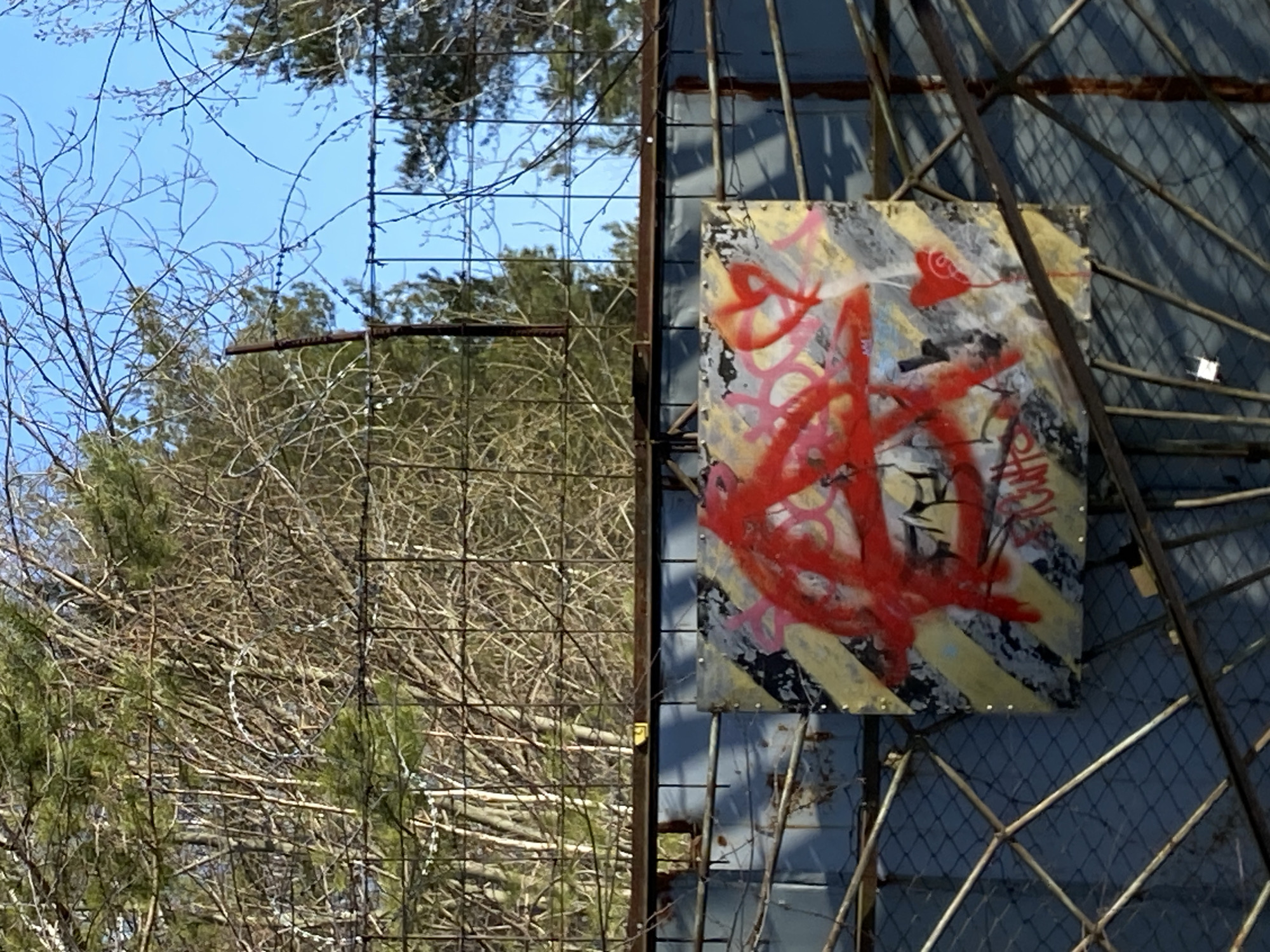 A medium, outdoor shot depicts a graffiti-covered, warning-striped sign affixed to a metal structure, situated near bare trees and a barbed-wire fence. The sign's primary colors include gray, yellow, and red, suggesting a possible urban or industrial setting. The presence of trees, a barbed-wire fence, and the weathered metal structure indicate an abandoned or overlooked location. Without any additional information in the prompt, this scene is difficult to conclusively identify it as a location in Chaikovsky, Russia.