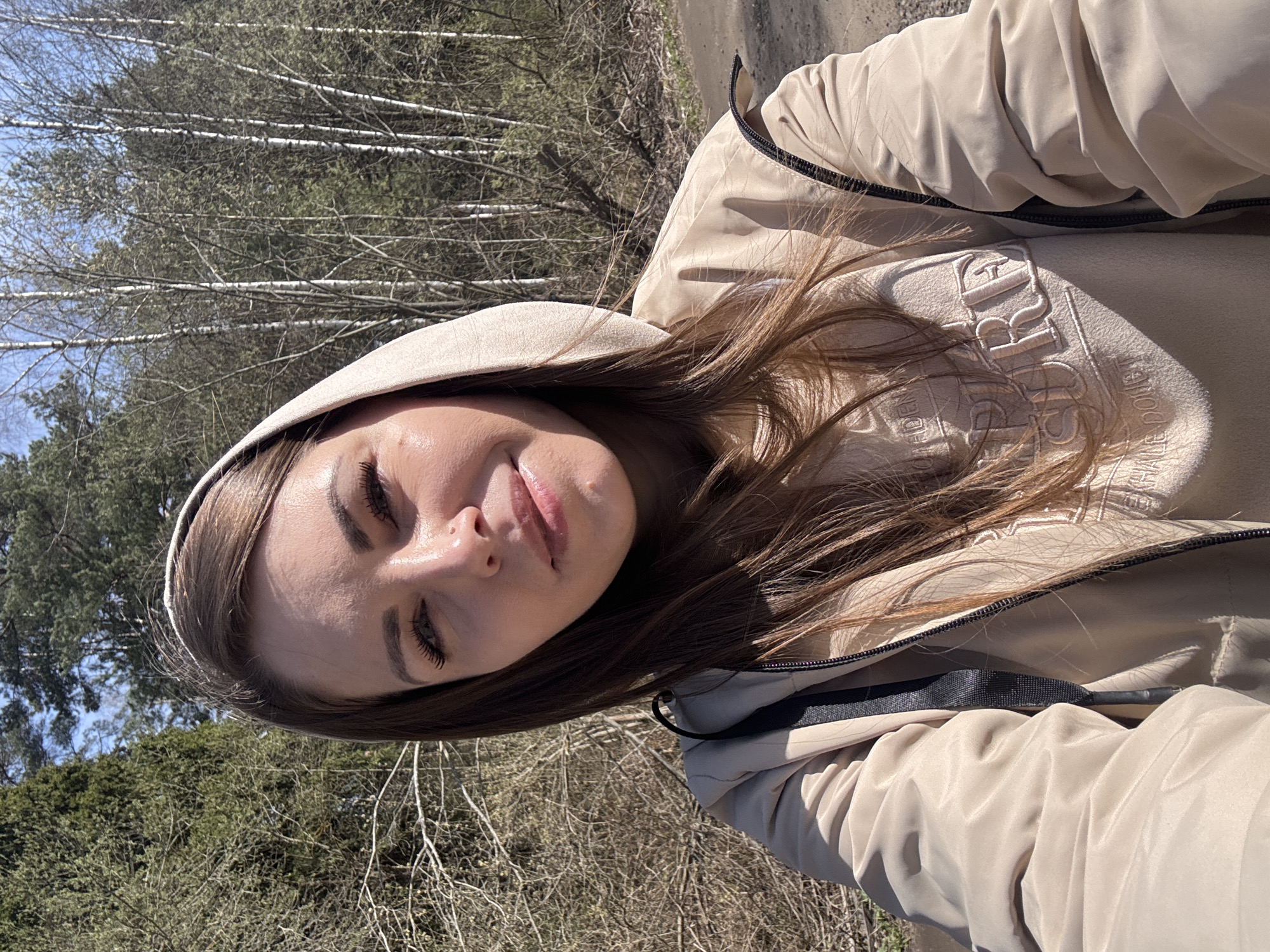 The image is a selfie of a young woman taken outdoors in Раздолье, Russia. She is depicted from the chest up, with her head tilted, looking towards the camera with a relaxed expression, her eyes partially closed and a subtle smile on her lips. She has long, light brown hair, and a white headband is visible on her head.
She is dressed in warm, casual attire, consisting of a light beige or cream-colored hooded sweatshirt and a matching beige jacket worn over it. On the chest of her sweatshirt, there is an embossed or embroidered design with partially visible text. The discernible parts appear to read "TRIS" followed by more letters, and below it, "GARDEN" and "MALE POLET."
The background suggests a natural, wooded environment. Several trees with distinct white bark, characteristic of birch trees, are prominent, appearing somewhat bare, indicating early spring or late winter. Other trees with green foliage are also visible, hinting at evergreens or trees beginning to leaf out. A clear blue sky is visible in the upper background. The scene is brightly illuminated by natural sunlight, suggesting a clear, sunny daytime. The overall ambiance is one of calm and natural beauty.