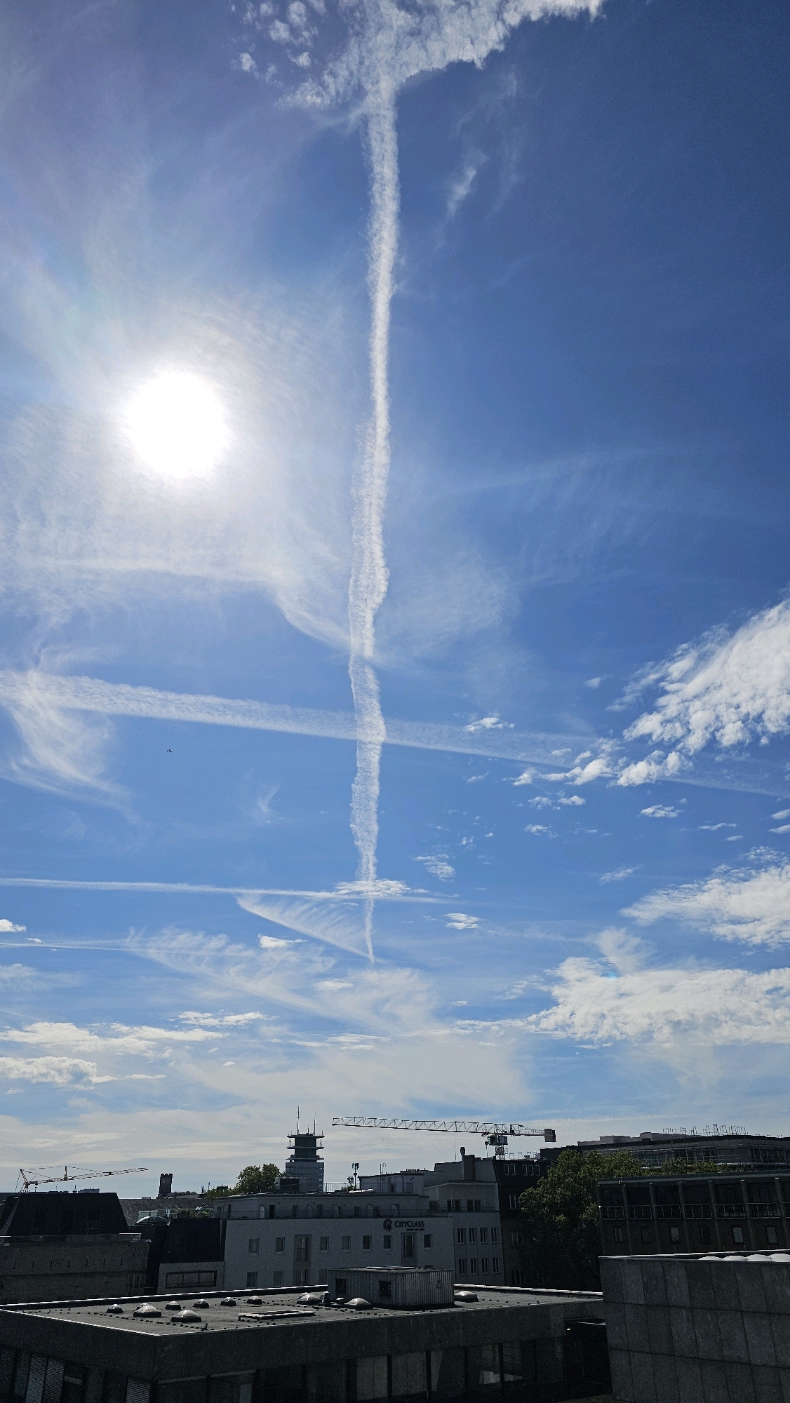 The image shows a bright, sunny day in Cologne, Germany. The sky is a clear blue, with several wispy white clouds and contrails from airplanes crisscrossing the sky. The sun is shining brightly, casting a glare on the lens. In the foreground, there is a rooftop view of several buildings, including some modern office buildings and older structures. Cranes are visible in the distance, suggesting ongoing construction. A small airplane can be seen in the sky. The scene is peaceful and urban, with a clear indication of a pleasant day.