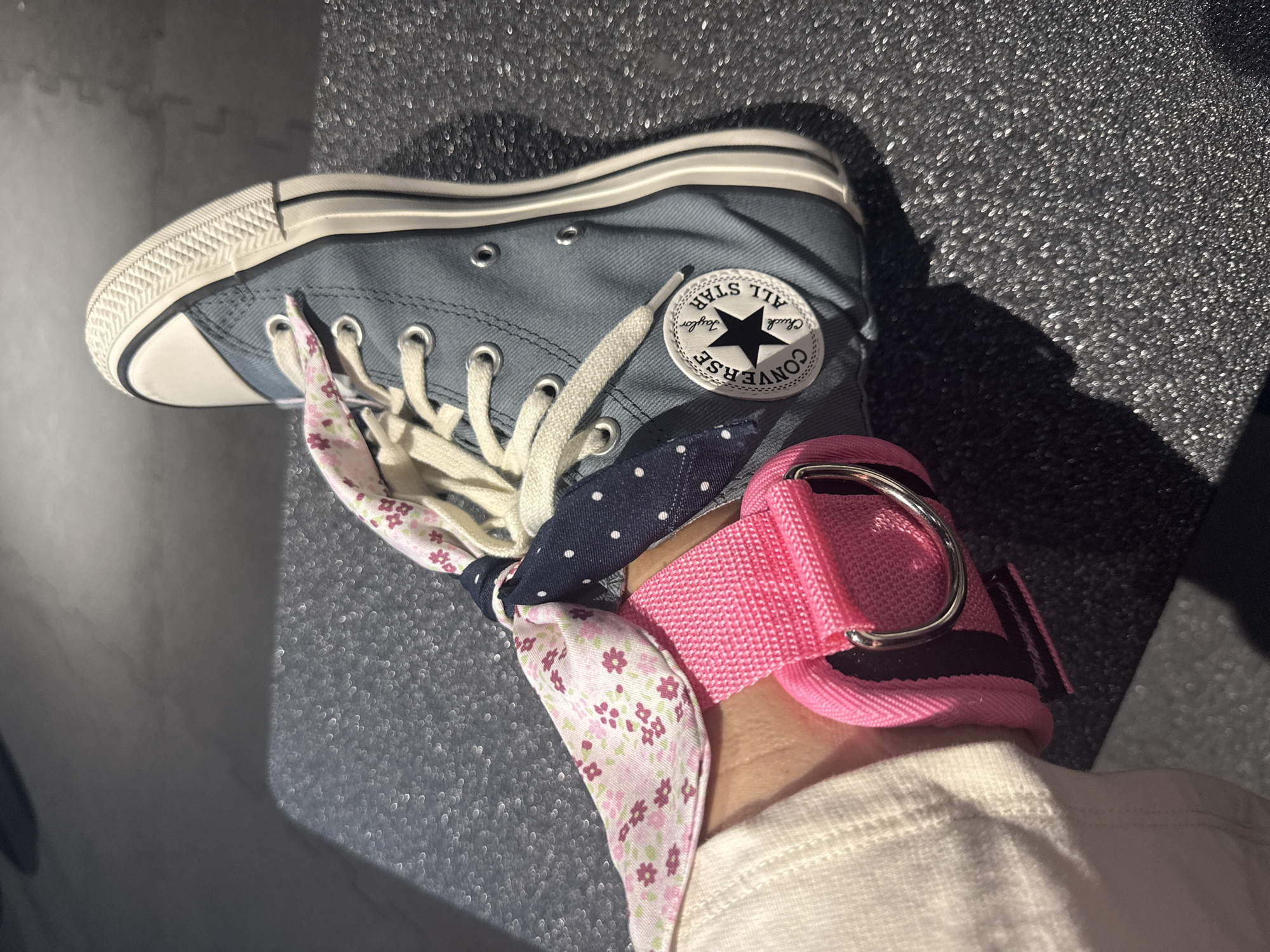 The image presents a high-angle, close-up view of a left foot adorned with a blue high-top Converse Chuck Taylor All Star sneaker, resting on a dark, granular-textured surface. The sneaker features white laces and the distinctive circular Converse All Star patch on the inner ankle, displaying the text "CONVERSE," "ALL STAR," and "Chuck Taylor" with a central black star.
Around the ankle, a personalized styling element is visible: two different patterned fabrics are intricately tied. One fabric is light-colored with a delicate pink and green floral pattern, while the other is dark blue with white polka dots, adding a playful contrast. Above these tied fabrics, a vibrant pink strap, possibly an ankle accessory or a component of exercise equipment, is fastened, featuring a prominent silver D-ring buckle. A sliver of a light-colored garment, likely a sock or pant leg, is visible beneath the strap.
Strong shadows are cast on the textured background, suggesting a bright, directional light source, indicative of daytime. The focus is on the detailed styling of the footwear and ankle. The image is contextualized as being taken in Valencia, Spain.