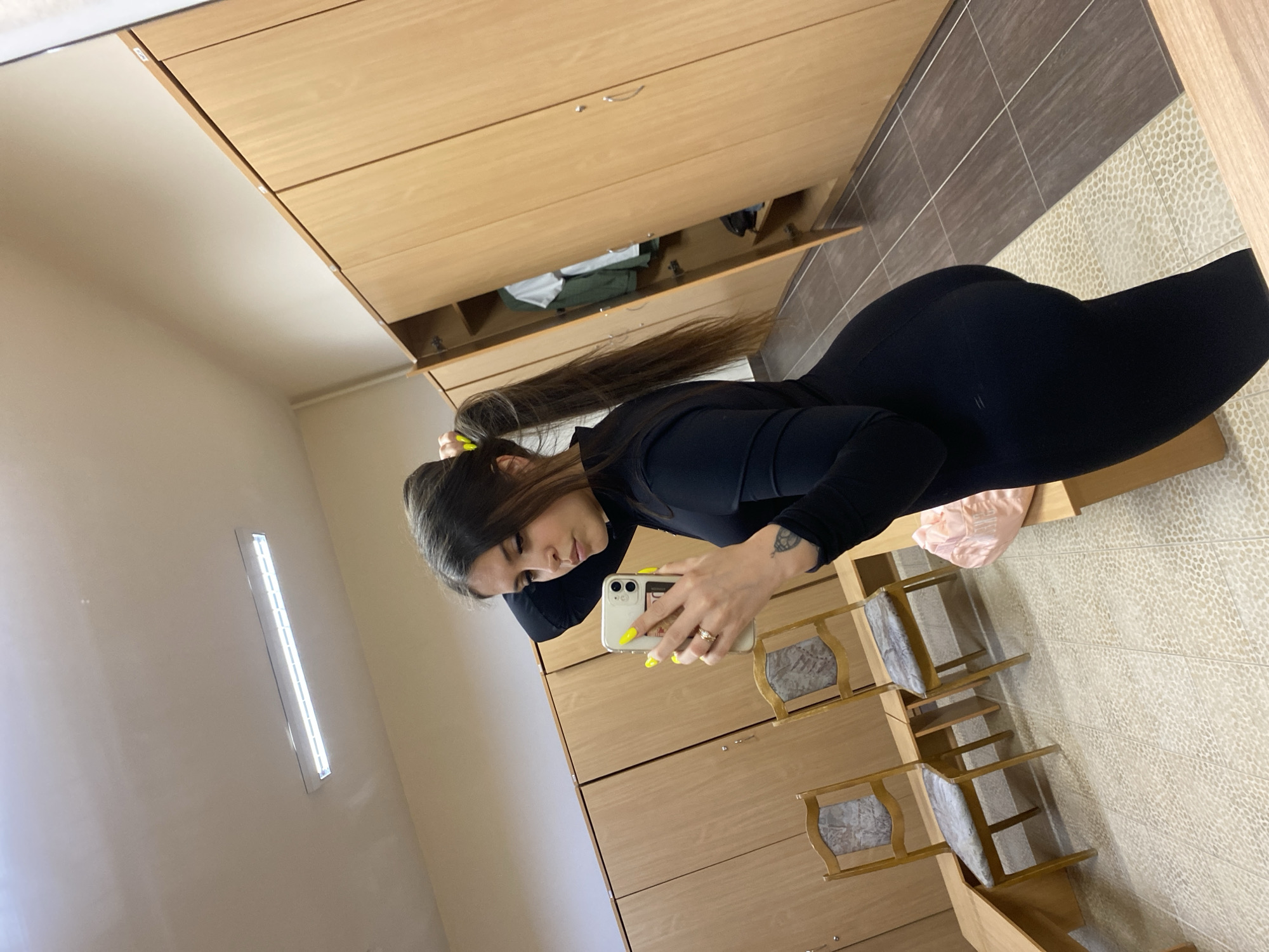 A woman is captured in a mirror selfie, standing in an indoor setting that appears to be a changing room or locker room. She is positioned to the right of the frame, facing partially away from the camera but looking down at her smartphone held up to take the picture. She has long, dark brown hair tied back with a bright yellow scrunchie or hair tie, and her face is visible as she focuses on her phone. She is wearing a black long-sleeved top and black form-fitting bottoms, possibly a dress or a jumpsuit. Her fingernails are painted a striking bright yellow, and she has a ring on her left ring finger and a tattoo on her left forearm.
The background, visible both directly and in the mirror reflection, features light-colored wooden cabinets or lockers with silver handles. Some of these cabinets are open, revealing folded clothes in green and white inside. A pink fabric item, possibly a bag or clothing, rests on a low wooden bench beneath the lockers. To the right, two wooden chairs with patterned, possibly grey, upholstered seats are arranged. The floor features a mosaic-like pattern in a light color, transitioning to darker grey/brown tiles in the reflection behind the woman. A long, rectangular fluorescent light fixture is visible on the wall in the reflection. The overall setting suggests an interior space like a gym changing area or similar facility. There is no visible text.