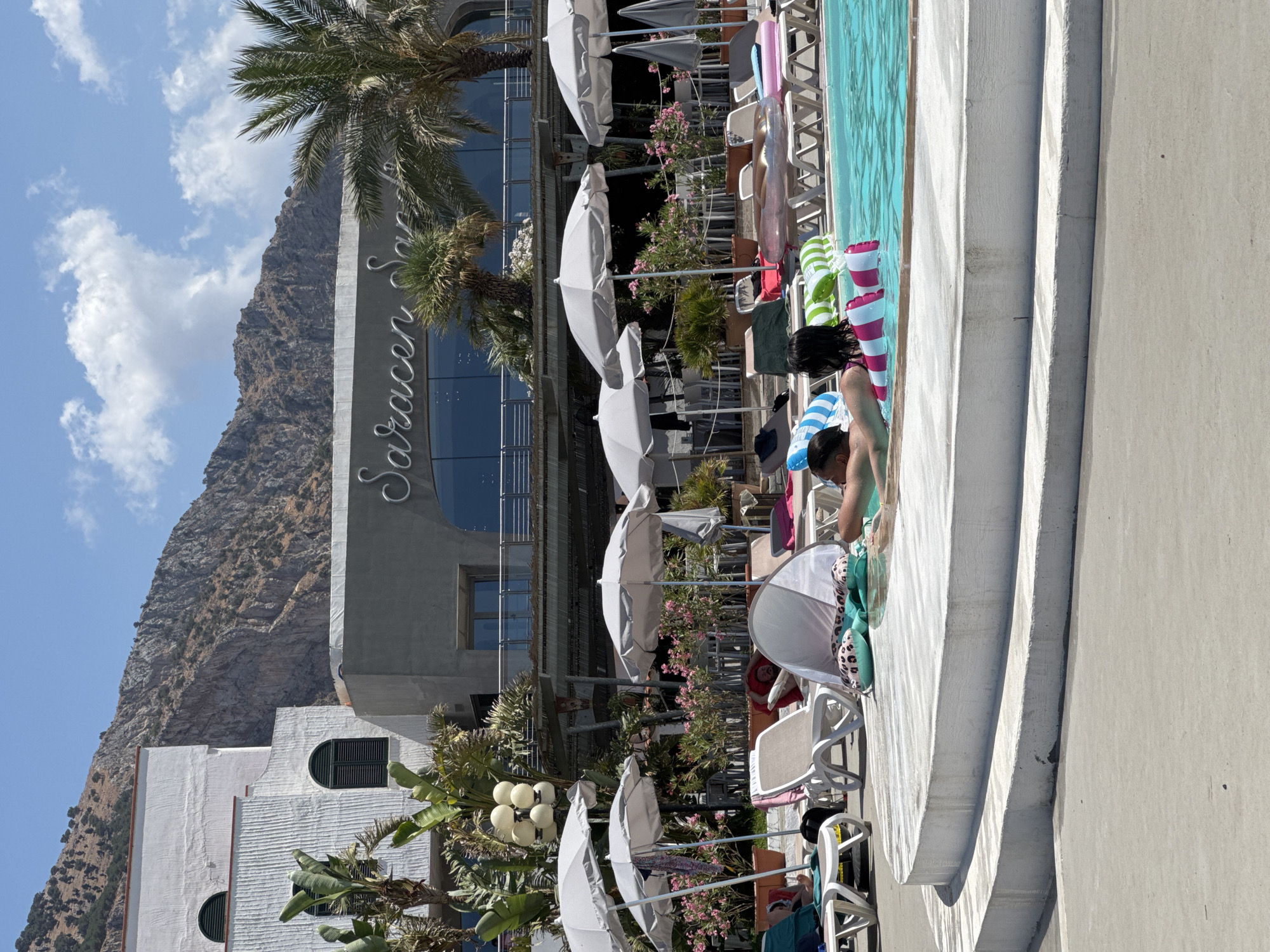 The image depicts a sunny day at a resort swimming pool located in Isola delle Femmine, Italy. In the foreground, two individuals are partially submerged in the bright blue water, relaxing on inflatable pool toys. One person has dark hair and is wearing a pink and white striped float, while the other person's face is partially visible. Surrounding the pool, white lounge chairs and several large, light-colored umbrellas provide shade. In the background, a modern building with the name "Saracen Sands" is visible, featuring large glass windows and a balcony. Palm trees and lush green plants add to the tropical ambiance. Behind the building, a rugged, mountainous landscape rises under a clear blue sky with scattered white clouds. The overall scene conveys a relaxed atmosphere typical of a vacation destination.