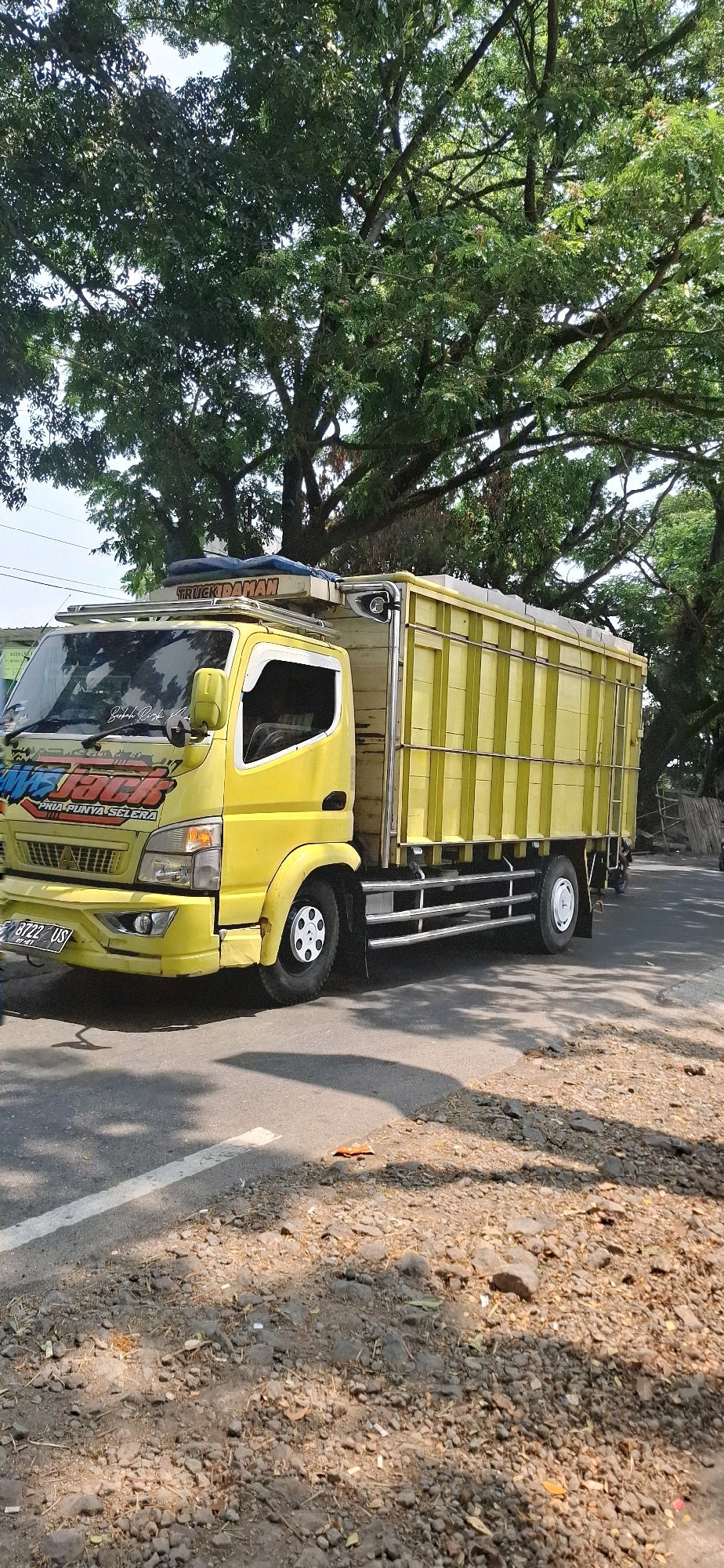 A yellow truck with a yellow cargo box is parked on the side of a road in Susukan, Indonesia. The truck has "TRUCK DAMAN" written on top of the cab and a sticker with "WES JACK" on the side of the cab. The license plate reads "B 8722 US". The truck is parked under a large tree with green leaves, and the ground is covered with rocks and debris. The sun is shining, casting shadows on the road.