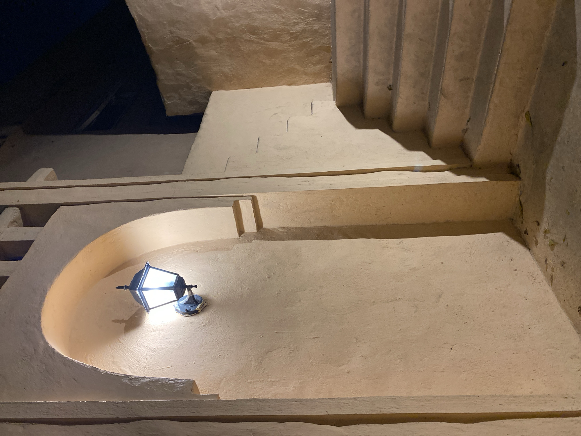 The image shows a lit outdoor lamp mounted on a textured, cream-colored wall beneath an arched opening. To the right of the lamp, a set of stairs ascends along another wall, also textured and cream-colored. The environment appears to be a courtyard or passage in an urban setting at night, with a dark sky visible in the upper right corner. There are no people or animals present. The overall impression is one of an architectural detail in a dimly lit area, possibly in a residential or commercial building complex. The location is identified as Đà Nẵng, Vietnam.