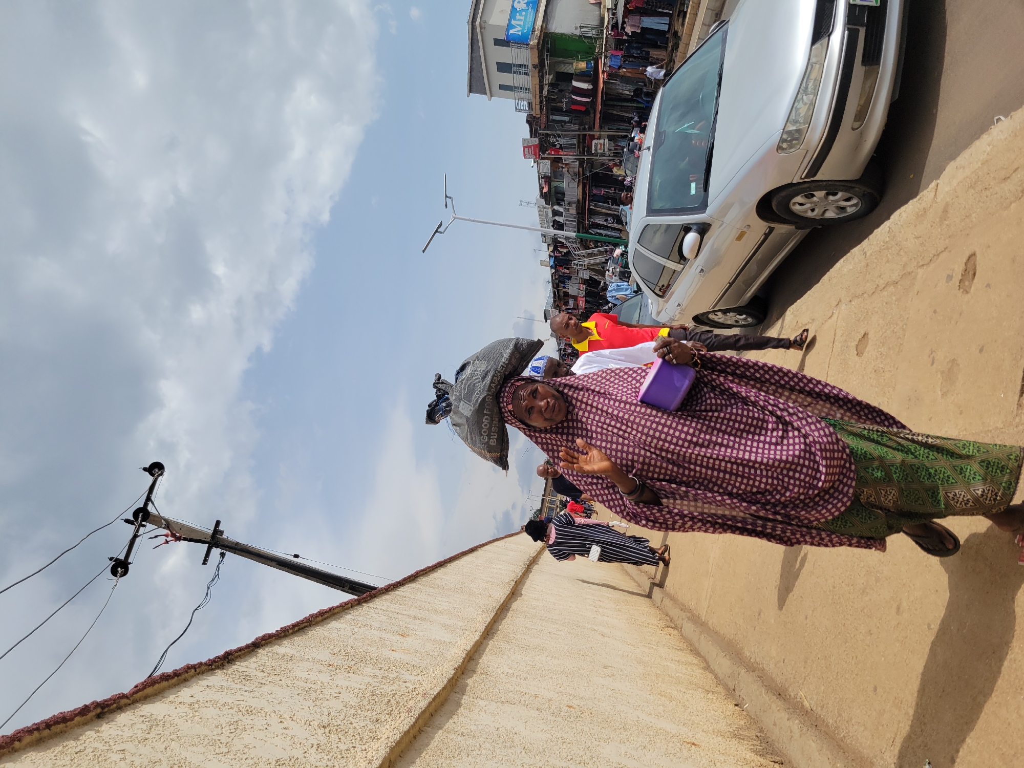 A street scene in an urban environment features a woman walking on a paved sidewalk carrying a large black bag of goods balanced on her head. She is wearing a patterned headscarf and traditional clothing, including a green patterned skirt, and is holding a purple object in her left hand. She waves with her right hand towards the camera. Several other people are visible walking behind her on the sidewalk, and a silver car is parked on the street to her right. The street is lined with buildings that appear to be shops with clothing displayed outside. Overhead, power lines are strung between poles against a cloudy sky. The ground is a light brown, and the wall on the left side of the image is a textured cream color with a red trim.