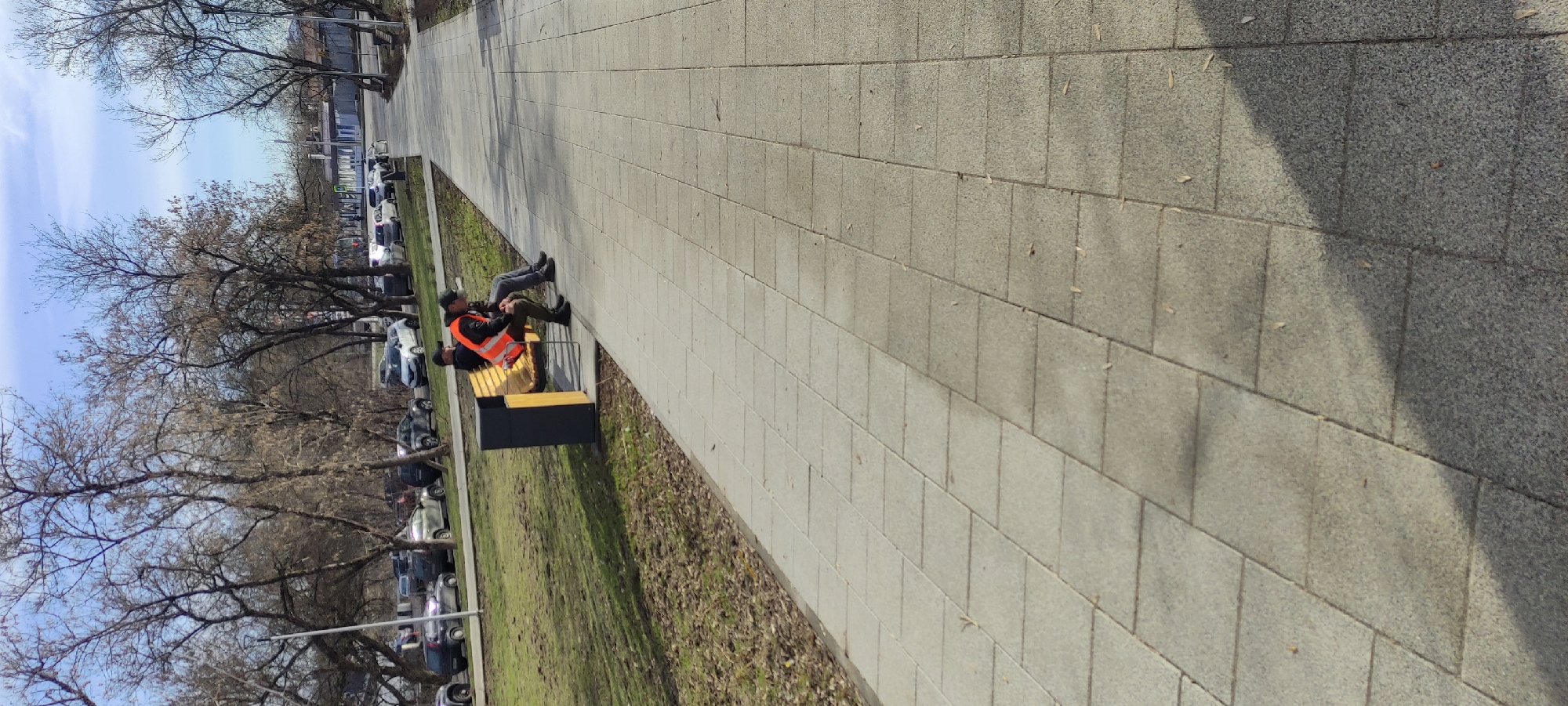 The image shows a paved walkway in Fryazino, Russia, with two people sitting on a bench. One person is wearing an orange vest. The walkway is lined with trees and parked cars. The sky is partly cloudy.