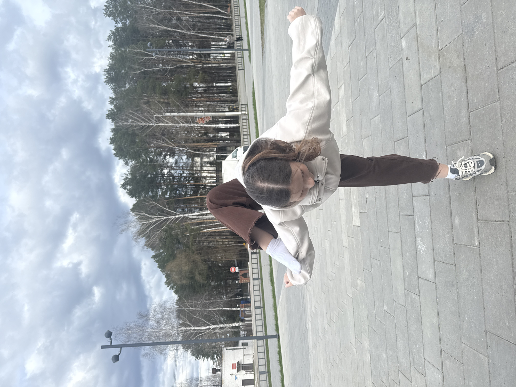 The image, taken in Chaikovsky, Russia, depicts a young person posing dynamically on a paved outdoor surface under an overcast sky. The individual, seen from above, wears a light-colored jacket or hoodie, dark brown pants, white socks, and grey and white athletic shoes. Their brown hair is visible, and they are striking a pose with one arm bent and raised, and the other bent and lower, conveying an active or playful stance.
The setting is an outdoor public space, possibly a park or wide sidewalk, featuring grey rectangular pavers. The background is dominated by a dense line of trees, comprising both tall coniferous species and bare deciduous trees, suggesting late winter or early spring. The sky is largely covered with grey clouds, indicating overcast daytime conditions and diffused lighting.
Notable elements in the background include a modern street lamp with two fixtures on the left, a low fence or barrier running along the tree line, and a red circular "No Entry" sign partially visible in the distance among the trees. The overall scene captures a moment of movement in an urban green space.