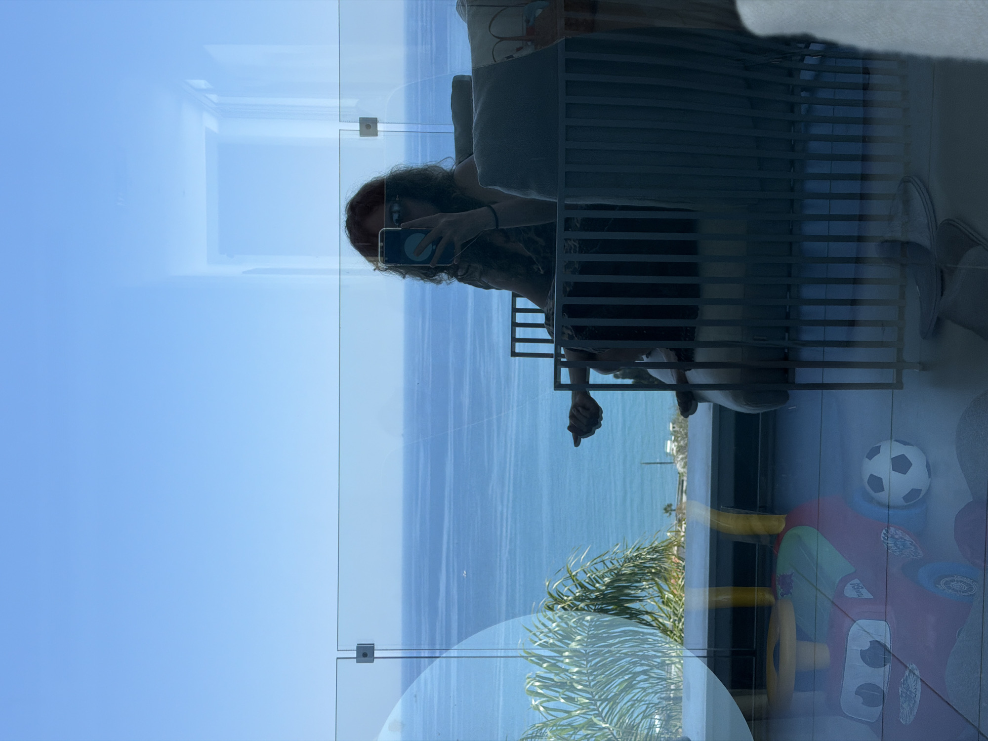 The image captures a reflection on a glass balcony overlooking the Mediterranean Sea. In the foreground, a red toy car with a soccer ball is visible on the tiled floor. A woman with curly red hair, wearing sunglasses and a patterned dress, is seated in a chair, holding a smartphone and taking a selfie. She is partially obscured by the glass railing and a metal barrier. In the background, the bright blue sea stretches to the horizon, with a small boat visible in the distance. Palm trees and a coastal landscape with buildings are also reflected in the glass. The scene appears to be set on a sunny day in Agios Tychona Community, Cyprus.