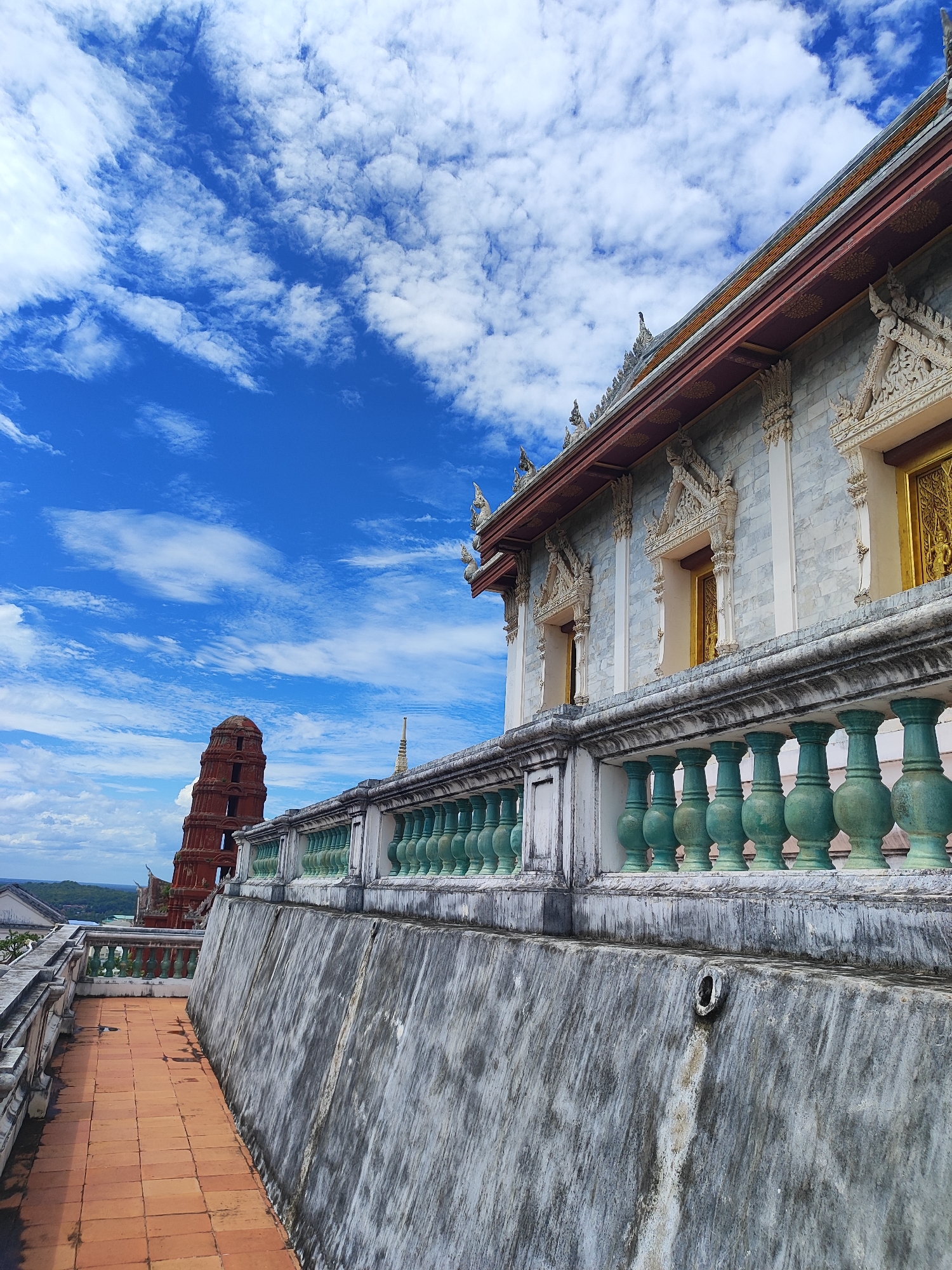 An outdoor, eye-level, full shot captures the upper section of a building in Phetchaburi, Thailand, with a terracotta tiled walkway in the foreground. The building, constructed of marble-like stone, features ornate golden doors and intricate carvings around its windows, with a decorative railing made of green balusters lining its roofline. In the distance, a reddish-brown, multi-tiered stupa stands against a vibrant blue sky dotted with white clouds. The scene appears to be part of a temple or historical site, likely Wat Phra Non.