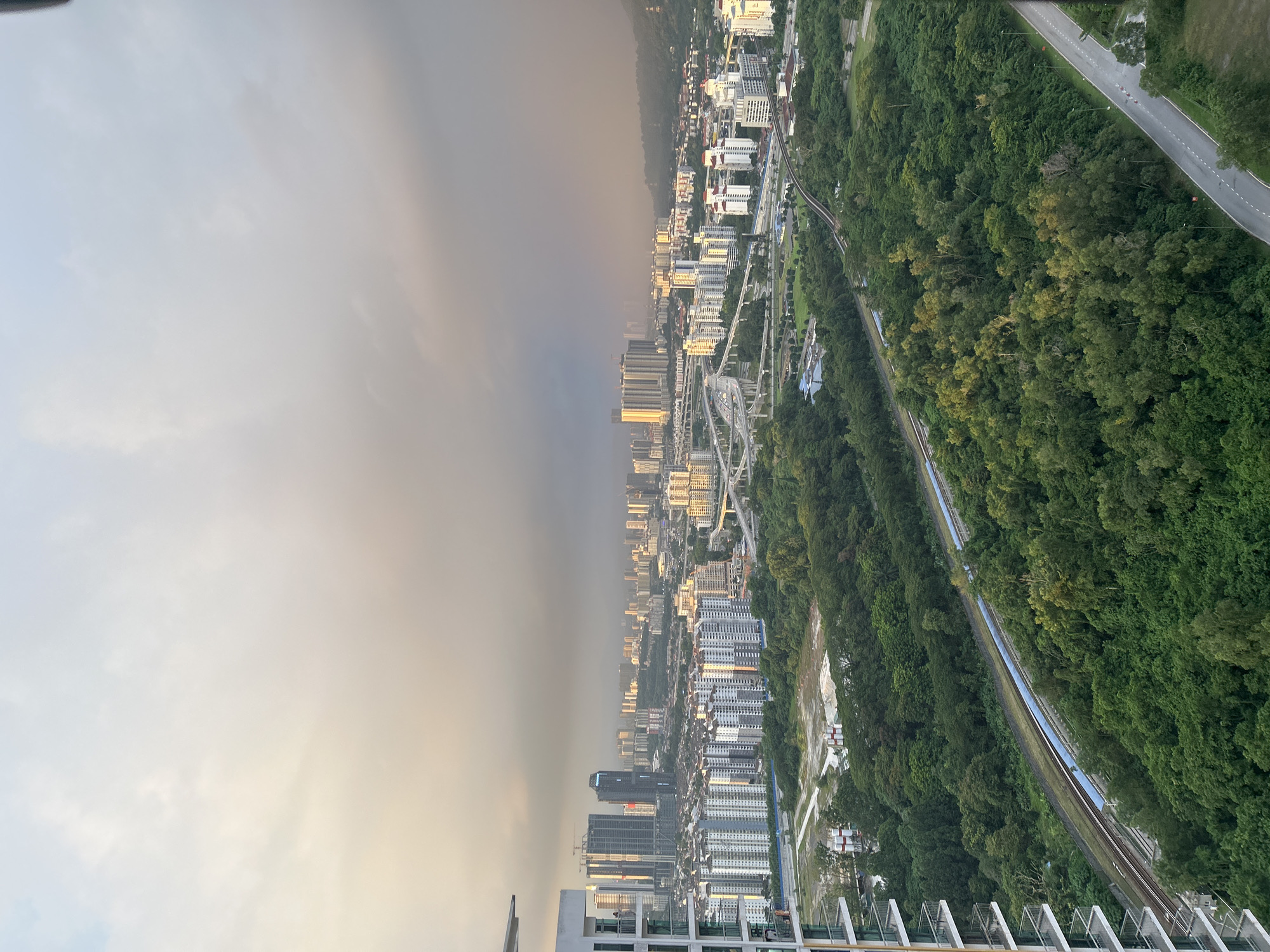 The image presents an elevated view of a landscape split between urban development and lush greenery, likely in Kuala Lumpur, Malaysia. On the left, a dense cityscape is visible, with numerous high-rise buildings reflecting sunlight. Construction cranes suggest ongoing development. The sky is overcast, with a visible demarcation between a clear sky and a hazy or smog-filled area. To the right, dense forest covers a substantial area, punctuated by linear clearings indicating roads or railway tracks. A winding road or railway curves through the forest. The composition suggests a transition between urban sprawl and natural landscape, a common feature of Kuala Lumpur.