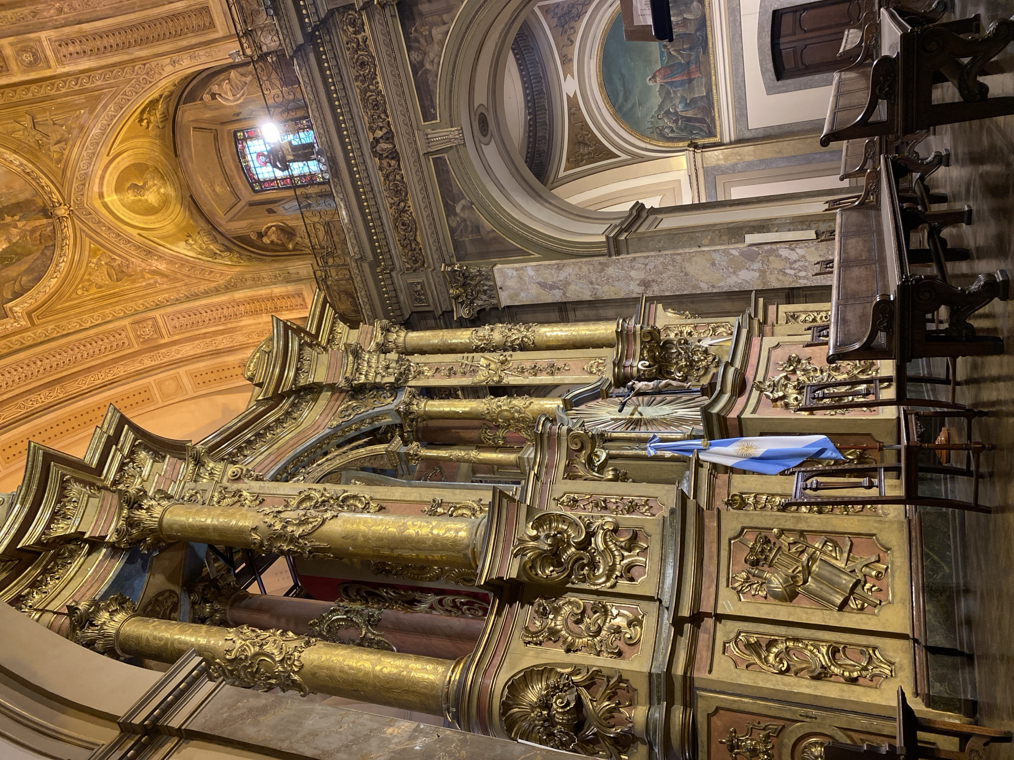 The image displays the opulent interior of a grand church or cathedral, likely located in Buenos Aires, Argentina, evidenced by a visible Argentinian flag. The scene is characterized by intricate, gilded architectural details and extensive religious artwork.
Dominating the view are massive columns and pilasters, richly adorned with gold leaf and elaborate carvings in a Baroque or Neoclassical style. These ornate structures support soaring arches and domed ceilings, which are covered in detailed fresco paintings or murals depicting religious narratives and figures. One section of the ceiling features a vibrant stained-glass window, emitting a bright light into the space.
In the mid-ground, a large, multi-tiered structure, possibly an altarpiece or reredos, stands prominently. It is heavily decorated with golden ornamentation and carved relief panels, one of which features a depiction of classical instruments or symbols. A blue and white Argentinian flag, bearing the Sun of May emblem, is draped over a part of this structure. To the right, several dark wooden pews or choir stalls with carved details are arranged. Higher up, within the painted dome, a figure on a cross is subtly visible.
The lighting within the church appears to be a mix of natural light filtering through the stained glass and artificial illumination, casting a warm, reverent glow over the gilded surfaces and artwork. The photograph is taken from a slightly low angle, highlighting the vastness and intricate details of the sacred space. No discernible text is visible in the image.