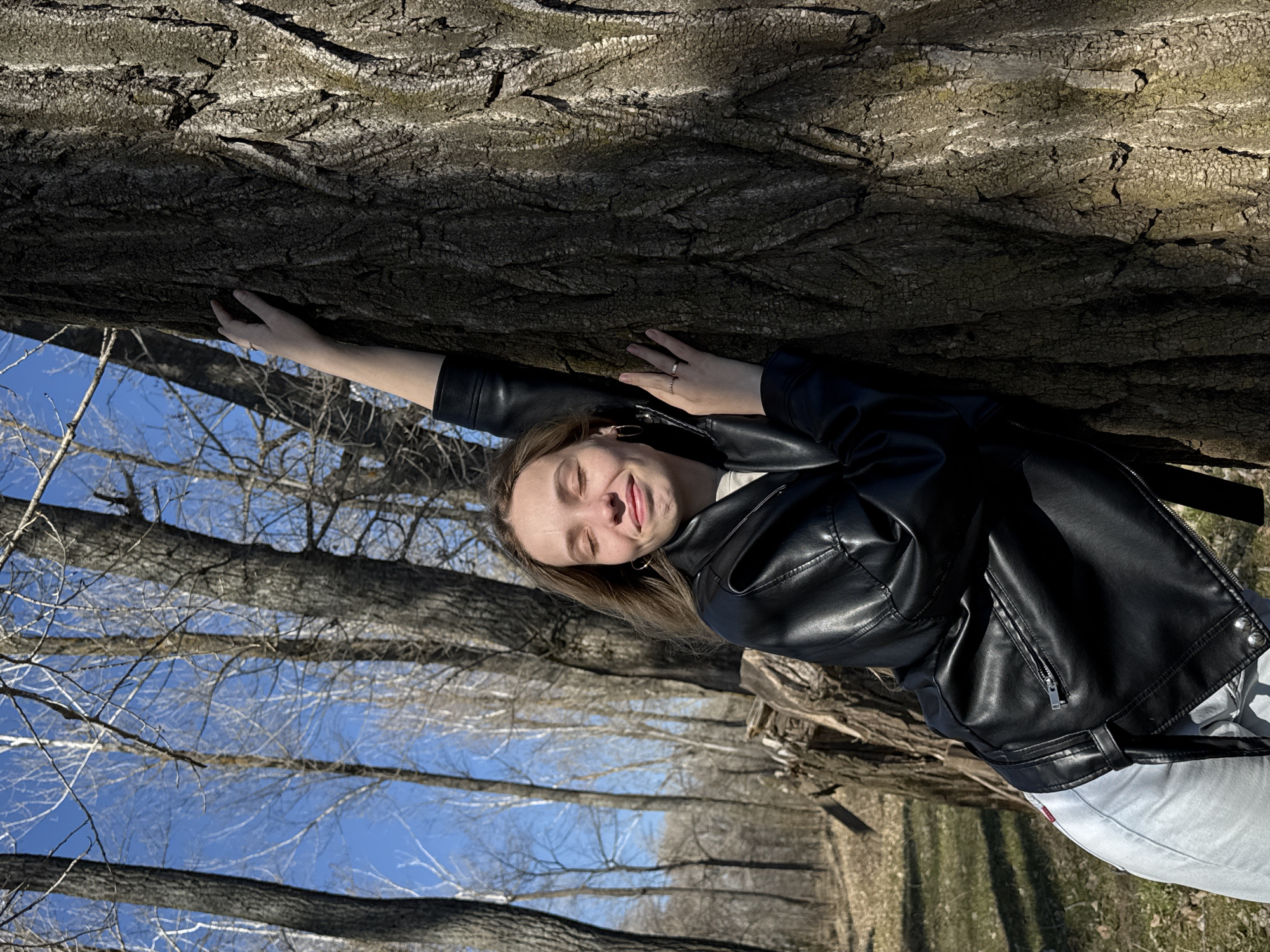 The image depicts a woman leaning against a tree in what appears to be a forest or park-like setting, possibly in Noviy, Russia. She is wearing a black leather jacket, light-colored jeans, and has her eyes closed with a serene expression. Her left arm is extended, touching the tree trunk. The tree has rough, textured bark. Behind the woman and the tree are other trees with bare branches against a blue sky. The ground appears to be covered in dry leaves or grass. The photo is oriented in a way that makes it seem as if the tree is lying horizontally.