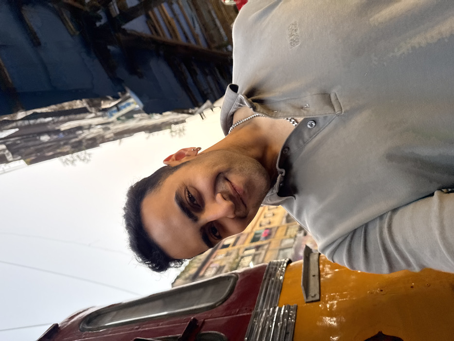 A male individual, appearing to be in his 20s or 30s, with dark hair and a light complexion, is depicted smiling directly at the camera. He wears a light gray long-sleeved polo shirt with a "BOSS Hugo Boss" logo embroidered on the left chest, a silver-colored cuban link chain necklace, and two silver-colored hoop earrings in his left ear. The subject's head is tilted slightly to the right.
To the immediate left of the individual, a partial view of a red and yellow public transport vehicle is visible, characterized by a red upper section, a yellow lower body, and horizontal metallic trim. This vehicle is identifiable as a Kolkata tram. In the background, an urban street scene unfolds under soft daylight conditions. On the left, behind the tram, a multi-story, aged residential or commercial building with numerous windows and balconies is visible, with clothes hanging on an upper balcony. To the right, behind the subject, a dense cluster of older, multi-story structures appears, exhibiting signs of wear and construction activity. The presence of the distinctive tram and the architectural style of the background buildings confirms the location as a street in Kolkata, India.