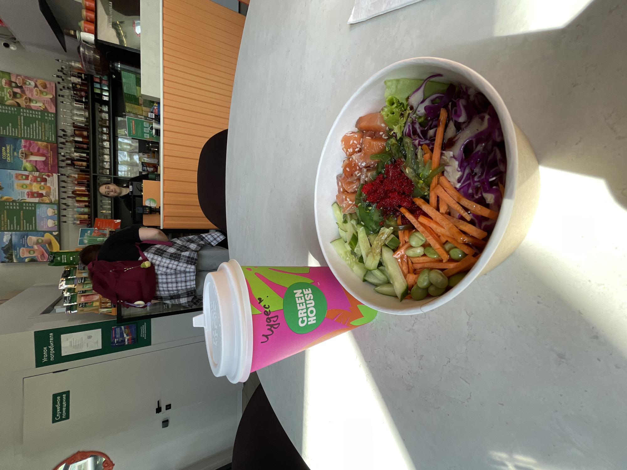The image, taken in Krasnoyarsk, Russia, depicts an indoor scene, likely a cafe or fast-casual restaurant, with a focus on food and drink on a table.
In the foreground, a large poke bowl is prominently displayed on a light-colored, possibly stone or laminate, tabletop. The bowl is filled with various ingredients, including diced salmon, bright red caviar (likely tobiko), green seaweed salad (wakame), julienned carrots, sliced cucumbers, edamame beans, shredded red cabbage, and lettuce. The salmon pieces are sprinkled with sesame seeds. Next to the bowl is a tall, bright pink and green cup with a white lid. The cup features the logo "GREEN HOUSE" and what appears to be handwritten text in Cyrillic, "Чудес" (Chudes, meaning "Wonders" or "Miracles"). Sunlight streams in from the right, casting distinct shadows of objects and perhaps a window frame on the table.
In the background to the left, there's a service counter or bar area. Shelves are stocked with numerous bottles, and several menus or promotional posters, mostly in Russian, are visible on the wall behind the counter. One poster clearly reads "Собери свой микс" (Build your own mix). A staff member, a man with a beard, is visible behind the counter, looking towards the camera.
In the background to the right, another person, likely a customer, is seated. They are wearing a plaid shirt and have a red backpack on their lap. Further back, two white signs in Russian are affixed to a white wall or door: "Уголок потребителя" (Consumer's Corner) and "Служебное помещение" (Staff Room / Utility Room).
The scene suggests a relaxed daytime setting, with people enjoying or preparing to enjoy a meal. The presence of poke bowls and branded cups indicates a health-conscious or modern food establishment.