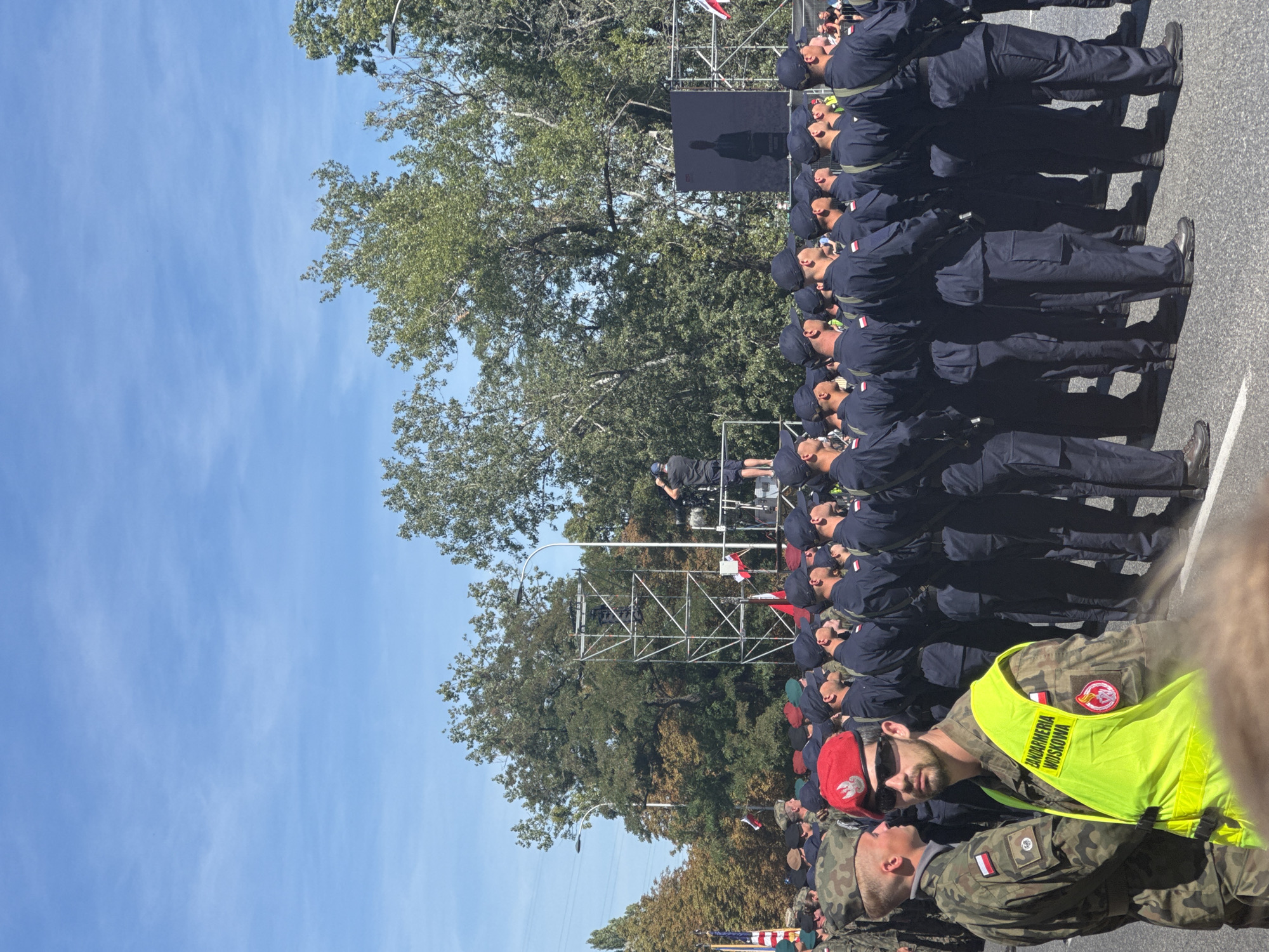 The image captures a daytime outdoor event, likely a parade or ceremony, in Warsaw, Poland. In the foreground, a man in a military uniform with a red beret and sunglasses is wearing a bright yellow high-visibility vest. The vest has text in Polish, including "ŻANDARMERIA WOJSKOWA," indicating he is part of the military police. Behind him, a formation of soldiers in dark uniforms and peaked caps stands at attention. Their uniforms appear to be dark blue or black.
In the middle ground, a large metal scaffolding structure is erected. A person is visible at the top of the scaffolding, appearing to film or photograph the event. Several Polish flags are displayed, some attached to poles and others incorporated into the scaffolding. A large screen displaying a silhouette of a person is also visible on the scaffolding. Trees with green and autumn foliage form the backdrop, suggesting the event is taking place in a park or a tree-lined street. The sky is clear blue with a few wispy clouds. The overall scene suggests a formal gathering with a military presence.