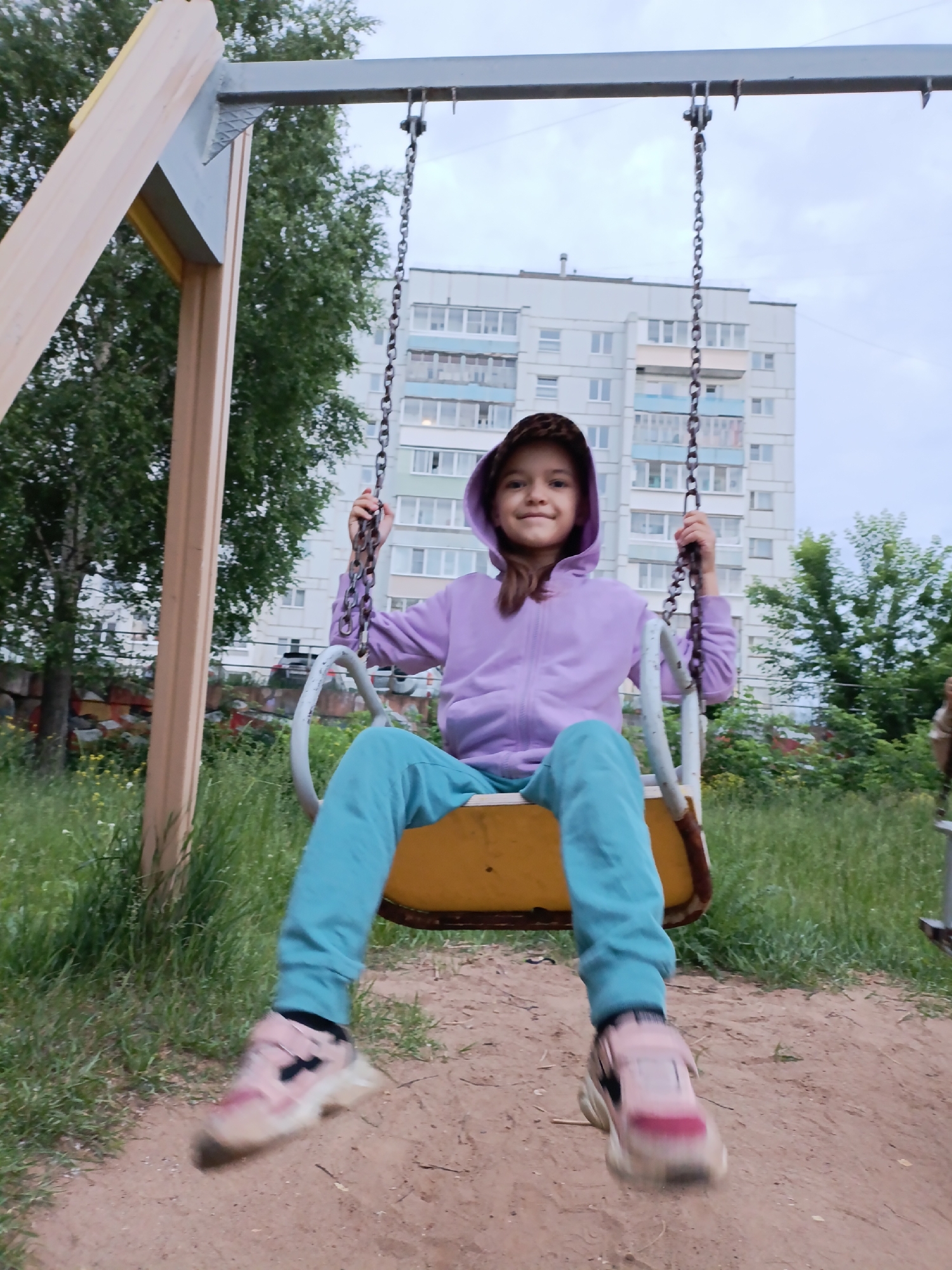 A young girl with brown hair, wearing a purple hooded sweatshirt and turquoise pants, is on a playground swing in a city park. She is holding onto the chains of the swing with her right hand and her left hand is partially visible. The swing seat is yellow with white accents. In the background, a tall, light-colored apartment building with many windows is visible, along with green trees and vegetation. The ground beneath the swing is made of sand and dirt. The scene is located in Chaikovsky, Russia, indicated by the architecture and overall setting. The lighting suggests it is daytime, possibly overcast.