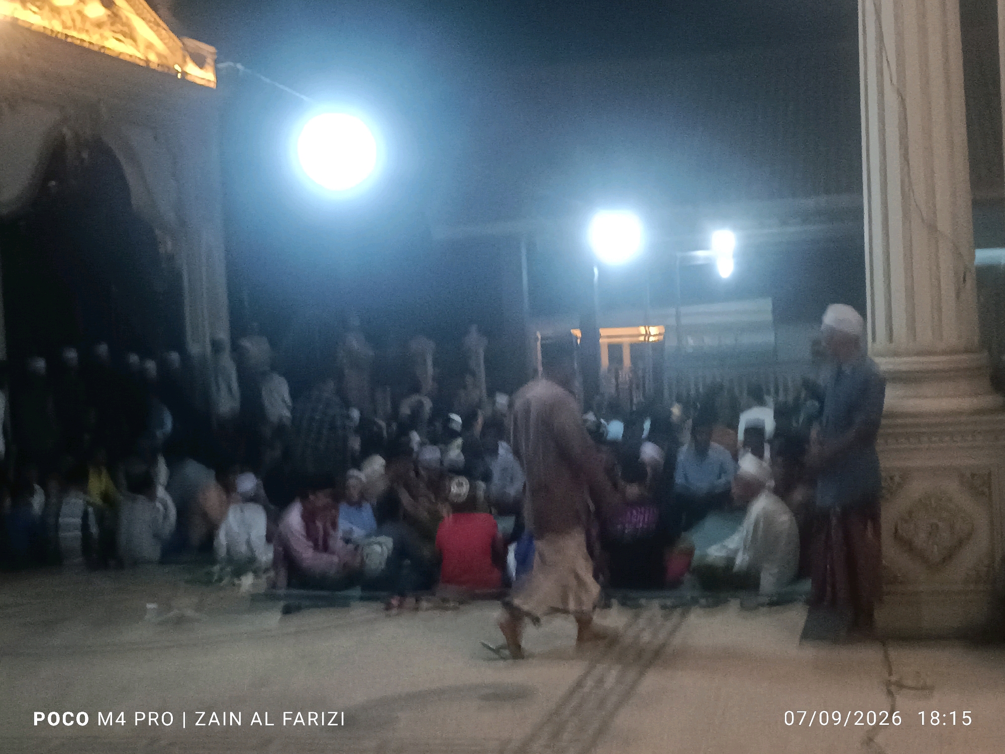 The image depicts an outdoor gathering of people at night, possibly at the Al-Aqobah Mosque in Buduran, Indonesia, given the metadata. The scene is dimly lit by artificial lights, creating a nocturnal atmosphere. A group of individuals are seated on the ground, appearing to be engaged in a communal activity or religious gathering. Some figures are standing, including a man in the foreground walking away from the camera. Architectural elements, such as a pillar and a building facade, are visible in the background, suggesting the location is a public space or a place of worship. The overall impression is one of a peaceful, late-evening assembly.