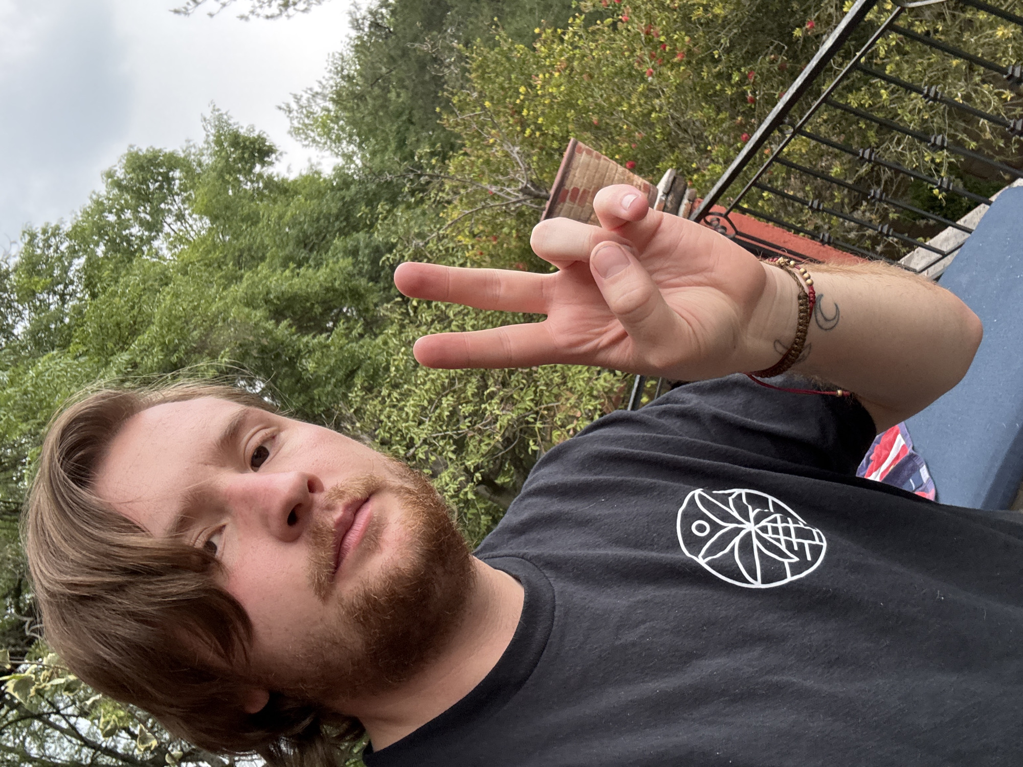A fair-skinned, bearded man with brown hair is positioned in the foreground, oriented at an angle, wearing a black t-shirt with a white circular logo featuring abstract designs. The man is holding up his hand, making a gesture with his fingers. On his wrist, a woven bracelet is visible, along with a tattoo. The background features greenery, suggesting an outdoor setting. A railing is partially visible in the upper right corner, alongside what looks like a wall or structure with red and grey elements. The environment includes trees and vegetation, and the sky appears overcast. Without further information or visual cues, it is impossible to locate the scene to Cañada de Madero, Mexico with any certainty.