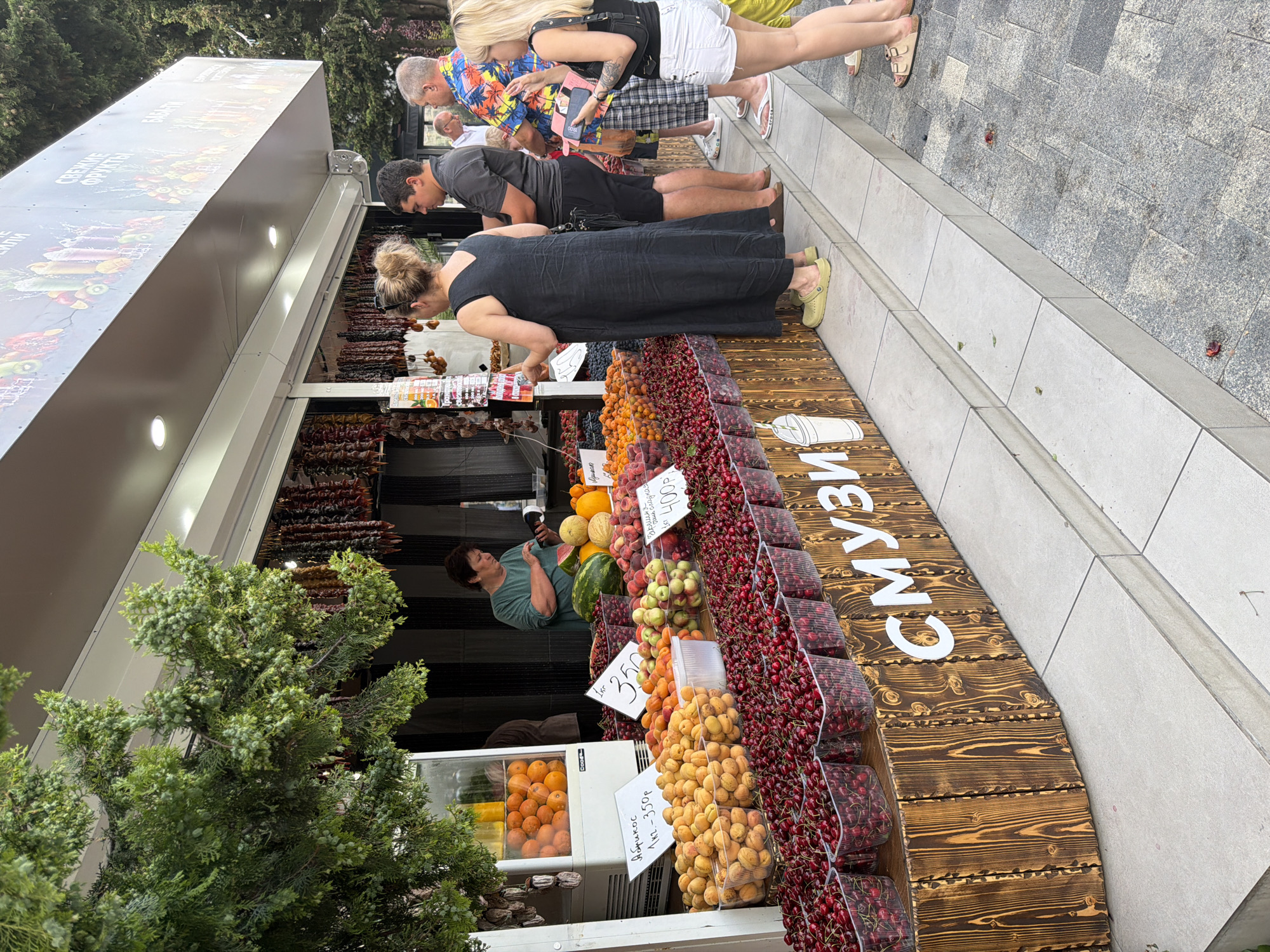 The image shows an outdoor fruit stand in Sochi, Russia. The stand is filled with various fruits displayed in clear plastic containers, including cherries, apricots, peaches, watermelons, melons, and oranges. There are also some dried fruit products hanging from the awning. Several people are gathered at the stand, appearing to be customers and a vendor. One woman, presumably the vendor, is seen behind the counter interacting with a customer, holding a device that may be a payment terminal. Other customers are looking at the fruit, some holding phones. The stand itself is made of wood with "СМУЗИ" (smoothie) written on the front in large white letters. In the background, there is a street or walkway with tiled pavement. The scene suggests a typical market environment in a city.