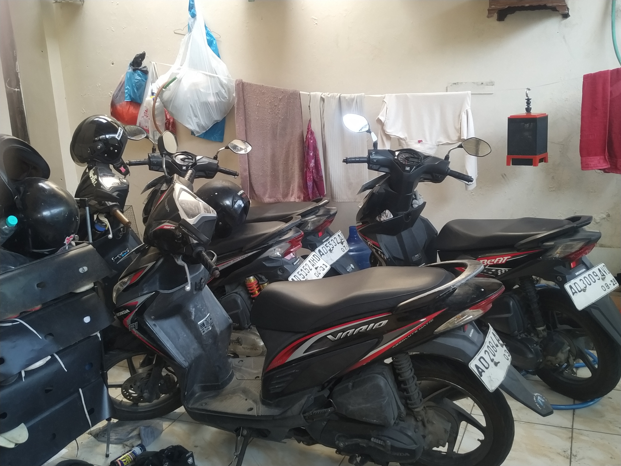 The image depicts an outdoor area with several parked motorcycles and laundry hanging on a clothesline. The motorcycles are of various models, predominantly black with red accents, and appear to be in a residential or a small business setting. Some motorcycles have helmets on them. The laundry includes towels and other garments, suggesting a domestic environment. There is a birdcage-like object hanging on the clothesline, with red and black detailing. The background consists of a textured wall. The scene is illuminated by natural light, casting some shadows. The location is identified as Banyudono, Indonesia.