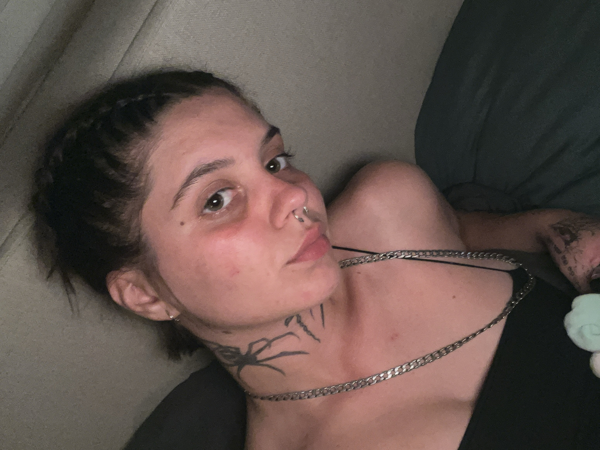 A young person with dark hair styled in a braid is lying down, facing the camera at an angle. They have a spider tattoo on their neck and a silver chain necklace. They are wearing a black tank top. The background appears to be a bed with a textured headboard. The lighting is dim, suggesting an indoor setting, possibly a bedroom. There is no indication of the city of Genemuiden, Netherlands.