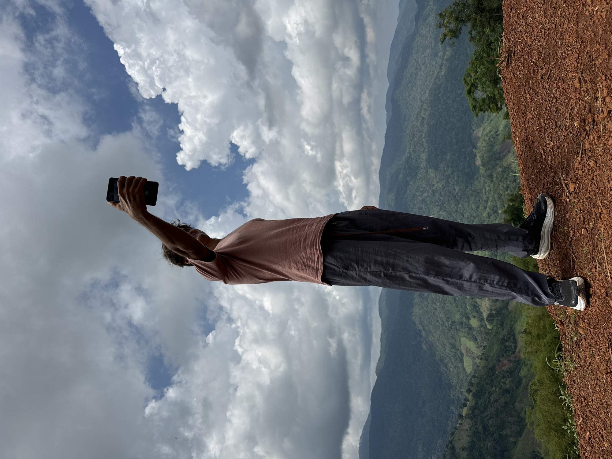 A person stands on a reddish-brown dirt ledge, holding a black smartphone aloft in their right hand to take a selfie. The individual is wearing a muted pink t-shirt, dark gray pants, and black athletic shoes with white soles. Their back is to the camera, and their head is turned slightly to the side, looking at the phone.
The background is dominated by a vast expanse of rolling, verdant mountains under a dramatic, cloudy sky. The clouds are a mix of white and dark gray, with patches of blue sky visible. The mountains are covered in dense forest, with some terraced fields or clearings visible on the slopes. The scene suggests an outdoor, possibly remote, location with a scenic mountain vista. The location is stated to be in Nao Sẻ, Vietnam.