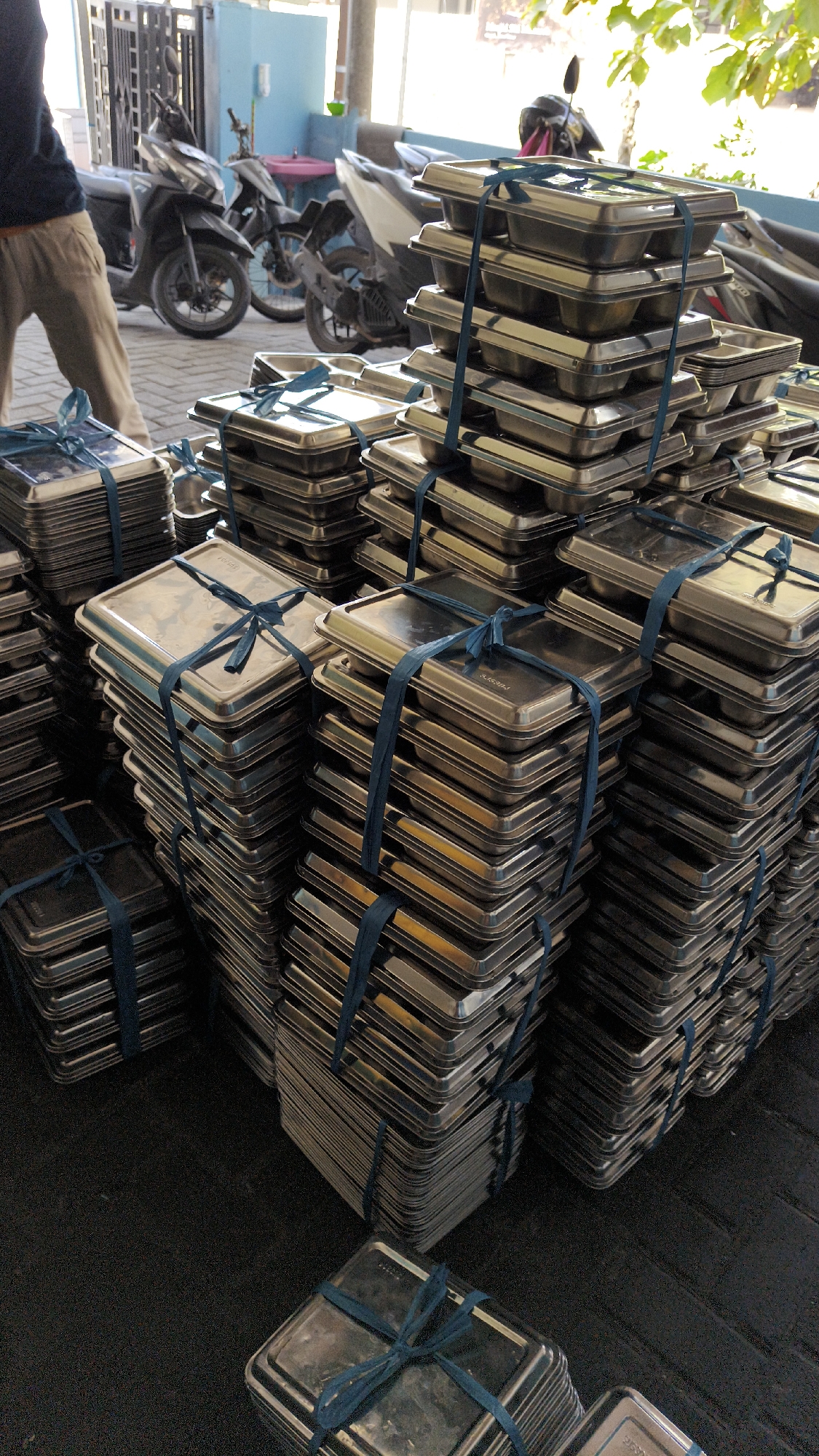 In Sambipondok, Indonesia, a person is seen from the waist down, wearing light brown pants and a dark blue long-sleeved shirt, walking on a paved surface. In the background, several motorcycles are parked. The foreground is dominated by large stacks of identical metal trays, neatly bound together with blue ribbon. These trays appear to be food containers, possibly for catering or distribution. Some stacks are quite tall, suggesting a significant quantity of these items. The environment appears to be an outdoor or semi-outdoor area, possibly a storage or distribution point. The lighting suggests it is daytime.