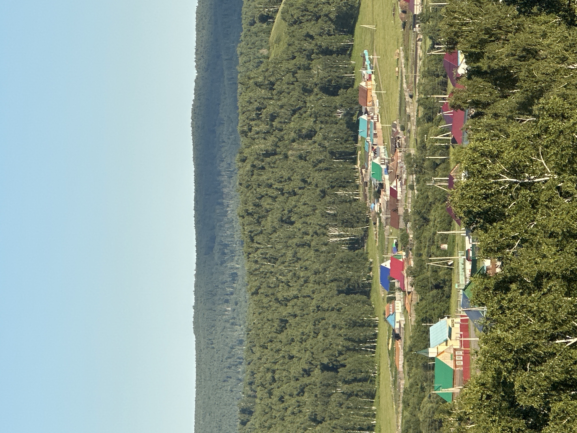 The image depicts a pastoral landscape with a village nestled in a valley. In the foreground, lush green trees, characteristic of birch trees with their white bark, dominate the view, their leaves creating a dense canopy. Beyond the trees, a village unfolds with a collection of houses, many featuring brightly colored roofs in shades of red, blue, and green, suggesting a vibrant community. The houses are of various sizes and architectural styles, with some appearing to be single-story residences and others possibly larger communal buildings. Some houses have red roofs and yellow or white walls, while others have green roofs and red trim. There are also some wooden fences and utility poles scattered throughout the village. In the midground, a vast, rolling hill covered in dense green forest, predominantly birch trees, rises majestically. The forest creates a textured backdrop, with lighter green patches suggesting clearings or lighter foliage. The background is characterized by a distant, hazy mountain range that appears as a darker, muted green silhouette against a clear, pale blue sky. The lighting suggests a bright, sunny day, likely in the late afternoon or early morning, given the long shadows cast by some of the structures and trees. The overall scene conveys a sense of rural tranquility and natural beauty.