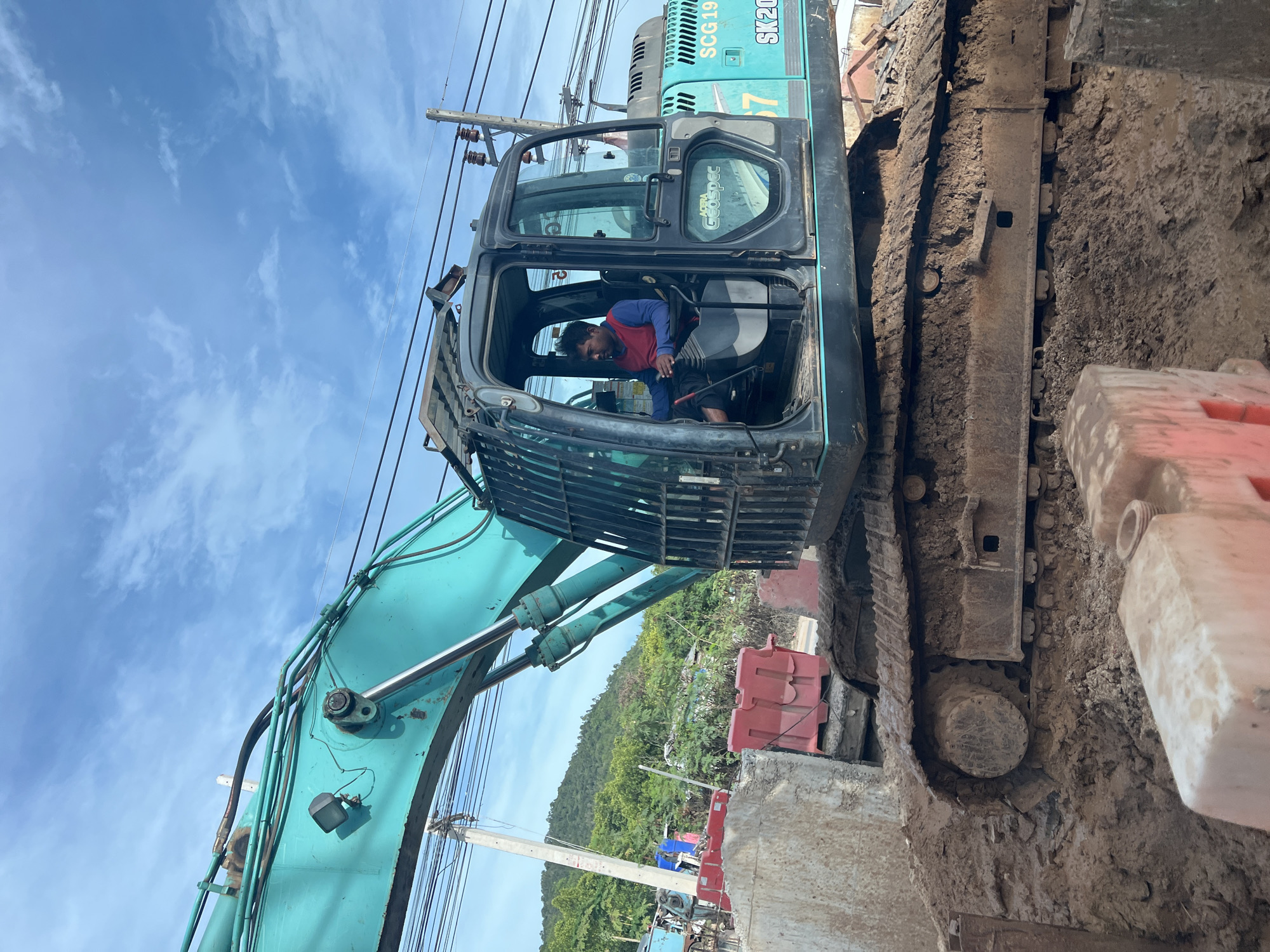A man in a red and blue shirt operates an excavator in Baan Bang Rak, Thailand. The excavator, bearing the model SK200, is positioned near a concrete structure bordered by red and white barriers. A utility pole with power lines is visible against a partly cloudy sky, with a forested hillside in the background. Mud is visible on the tracks of the excavator and on the ground.