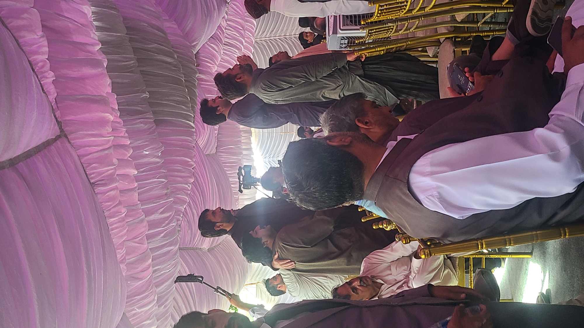 The image depicts a gathering of men inside a tent-like structure with white and pink draped fabric forming the ceiling. The men are seated in chairs, and some are wearing traditional attire like vests and shalwar kameez. Some appear to be using mobile phones. A video camera on a tripod is visible, suggesting the event is being recorded. The location is indicated as Quetta City Tehsil, Pakistan.