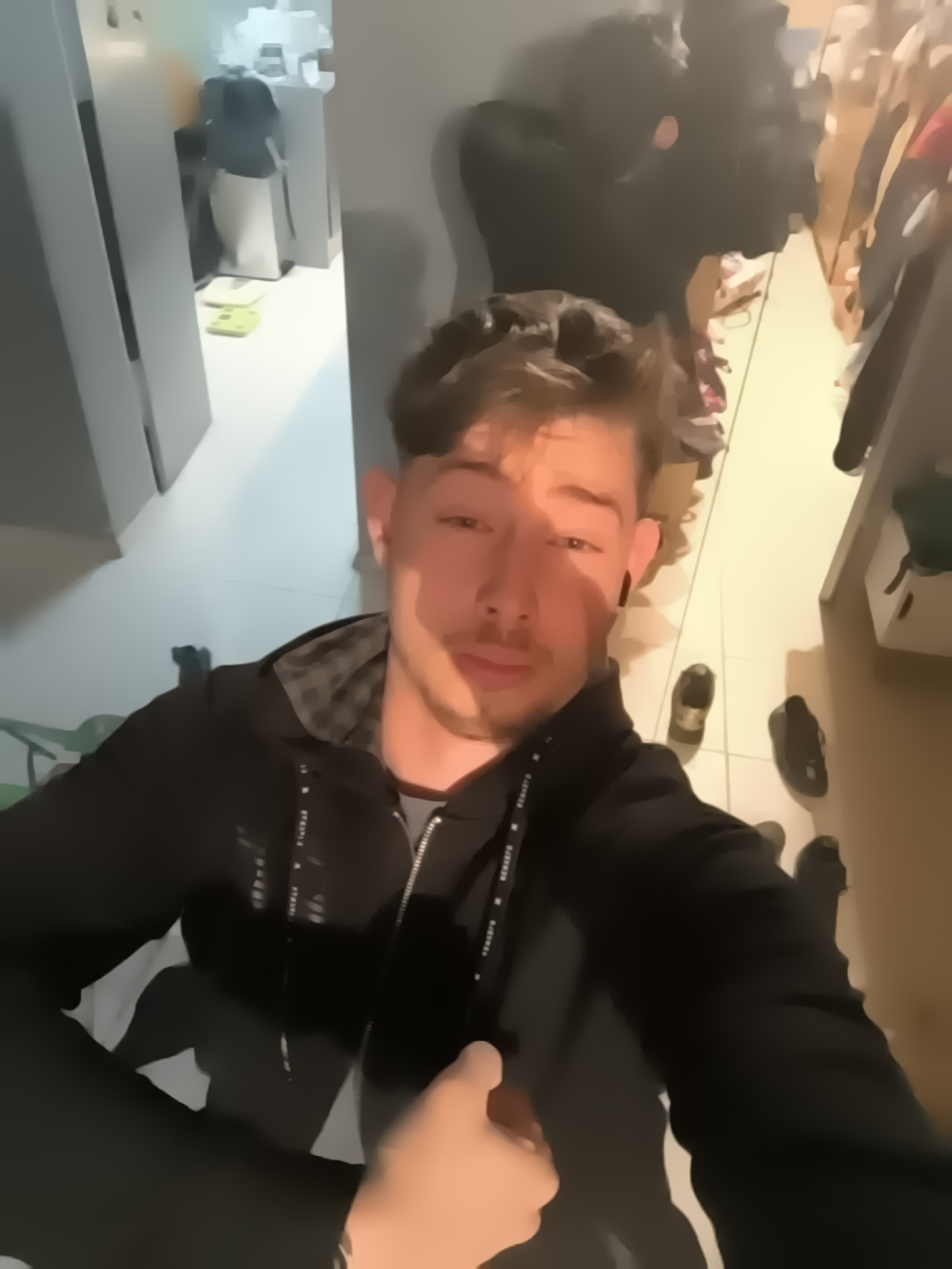 A person, likely male, is taking a selfie in what appears to be a hallway or entryway. He is wearing a black hooded jacket over a grey shirt. The hood has a checkered pattern lining. The hallway is cluttered with shoes and various items scattered on the floor. A closet or storage area is visible on the right side. On the left, there's a door or entrance with white cabinets. Based on visual context, the location in the city of Sofia, Bulgaria cannot be determined.