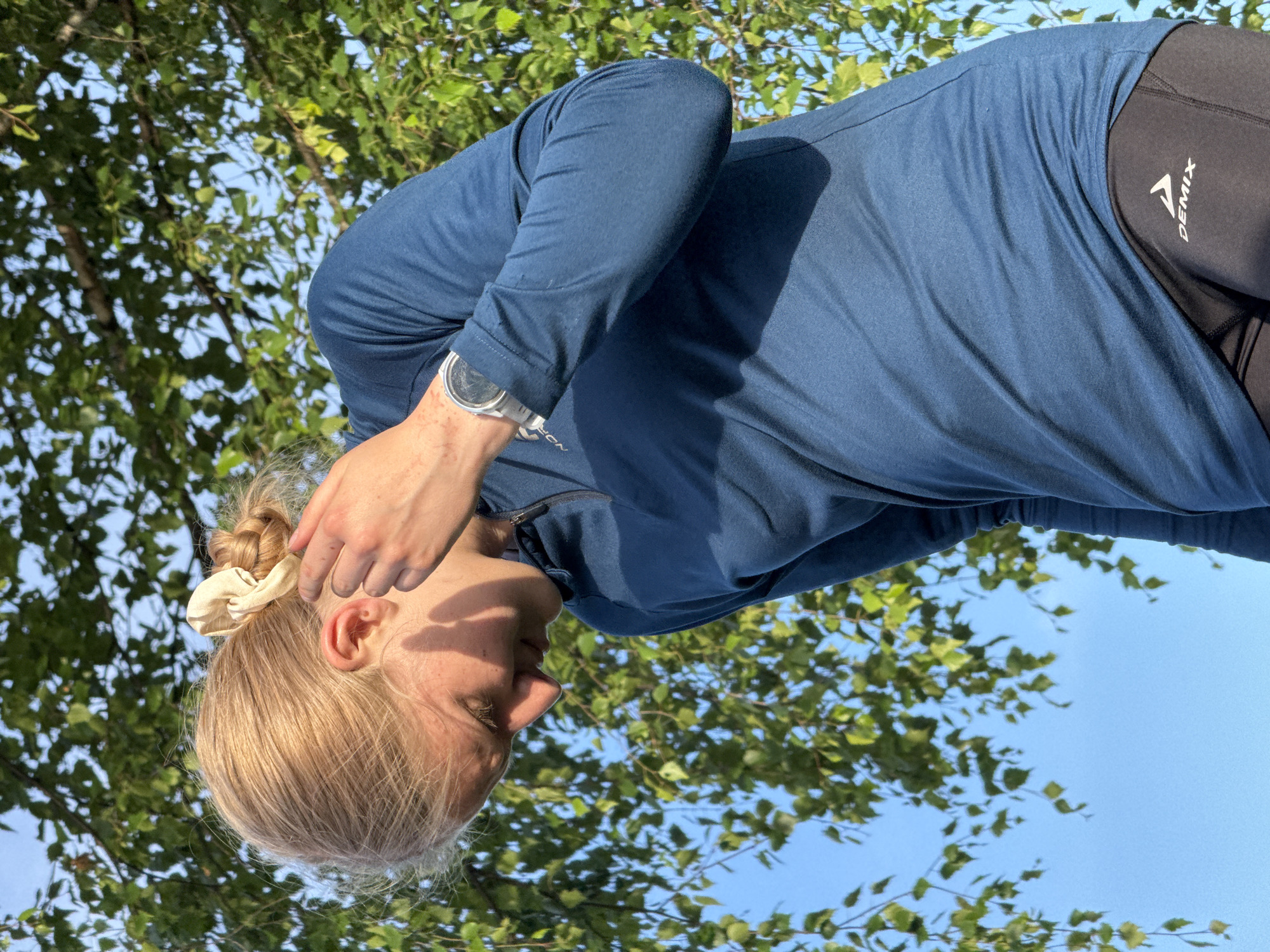 The image features a close-up, low-angle shot of a woman from the shoulders up, with her head tilted downwards. She has blonde hair pulled back into a bun with a light-colored scrunchie. Her right hand is touching her hair. She is wearing a dark blue long-sleeved shirt and black athletic leggings with a logo on the side that reads "DEMIX". A watch with a silver band is visible on her left wrist.
The scene appears to be outdoors, with lush green foliage and a clear blue sky in the background, suggesting it is daytime. The sunlight creates soft shadows on the woman's face and clothing. The overall mood seems relaxed and perhaps contemplative, with the woman possibly engaged in a moment of personal reflection or preparation for an activity. The setting, with the natural greenery and bright sky, suggests a park or a natural area.
The visible text is the brand name "DEMIX" on the woman's leggings. The image was taken in Sergino, Russia.