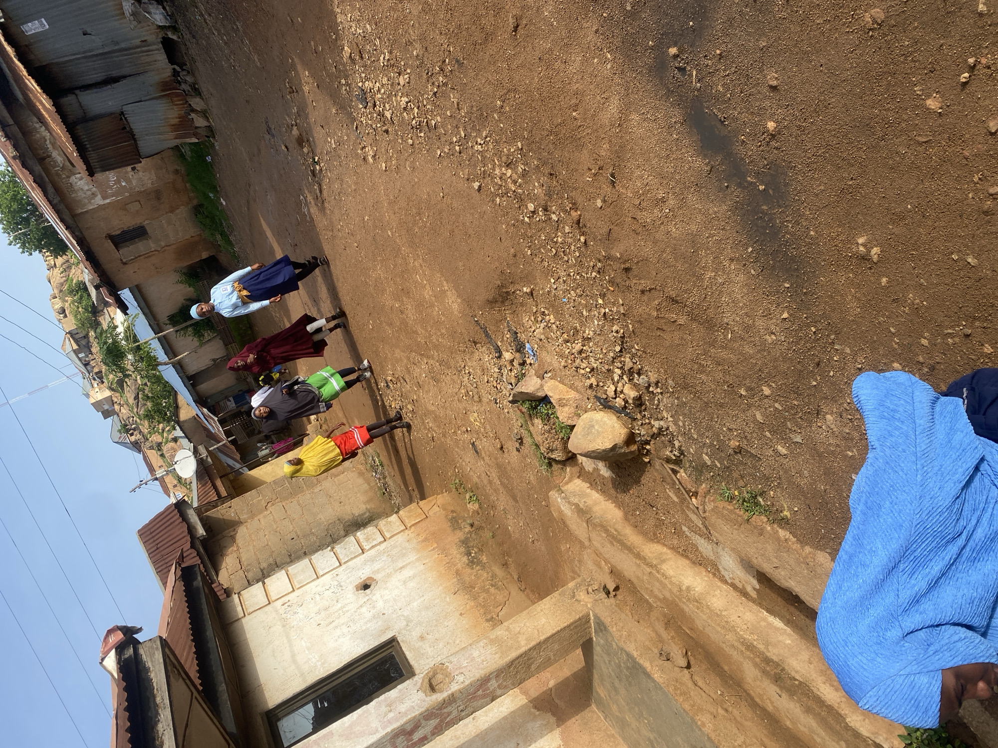 In Jos, Nigeria, a group of four young females, dressed in traditional Islamic attire, walk along a dusty, unpaved street. The buildings surrounding them are simple, brick structures with corrugated metal roofs, indicative of a developing urban area. One girl in the foreground wears a bright yellow hijab and a red skirt. To her left, another girl is in a black hijab and green skirt. Further down the street, a third girl in a maroon hijab walks with a fourth girl wearing a light blue hijab and a dark blue skirt. The sky is clear and blue, suggesting it is daytime, and the overall scene depicts a slice of daily life in this Nigerian city.