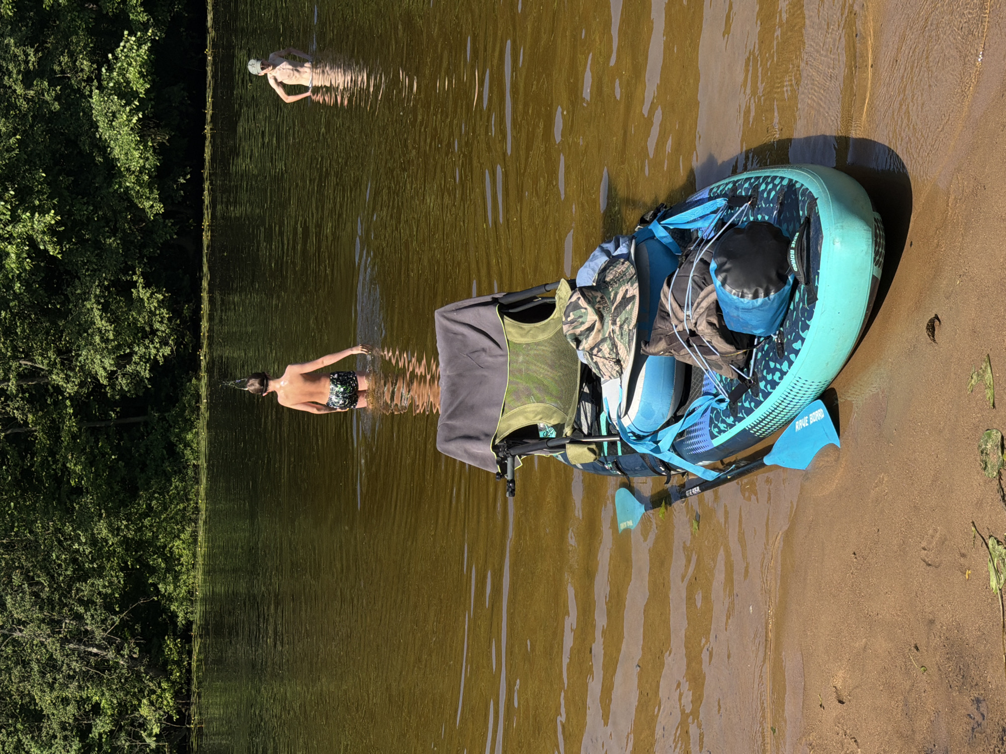 In Khimki, Russia, on a sunny day, a stand-up paddleboard is moored on the sandy shore of a body of water. The paddleboard is equipped with a portable chair and several bags, including a camouflage hat and a blue dry bag. Two young individuals are wading in the shallow water near the paddleboard. One, wearing patterned swim trunks, is further out in the water and looking away from the camera. Another person, wearing a cap and swim trunks, stands closer to the paddleboard, facing left. The background features a dense, green forest lining the opposite bank of the water.