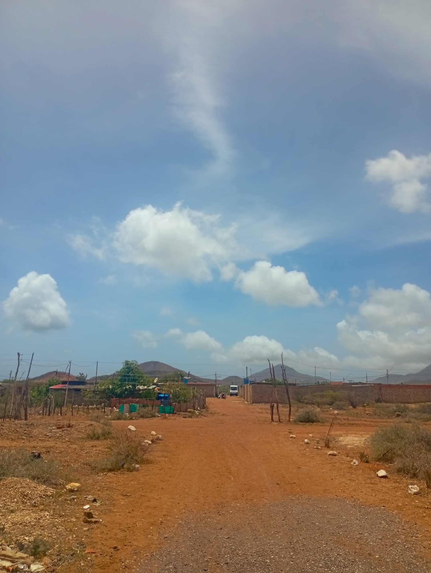 The image depicts an outdoor scene with a dirt road leading towards a cluster of buildings. The sky is partly cloudy with a bright blue hue. In the distance, mountains are visible. The foreground is dominated by the reddish-brown dirt road and dry, sparse vegetation. Several wooden posts and some fencing are present along the sides of the road.
A white van is parked on the left side of the road, near some low-lying buildings. To the right of the van, there appears to be a wall constructed of brick or concrete blocks. Scattered rocks and small bushes are visible on the ground.
The buildings in the middle ground seem to be of a simple construction, with some exhibiting corrugated metal roofs or tarps. Trees are present in this area, some of which are leafy and green, contrasting with the dry surroundings. There are also some objects that could be blue tarps or containers near the buildings.
The overall impression is of a somewhat arid or rural environment. Given the location specified as Altagracia, Venezuela, the scene is consistent with a rural or semi-urban setting in that region, characterized by its climate and typical infrastructure. There are no clearly identifiable individuals in the frame. The focus is on the landscape, the road, and the sparsely populated structures.
