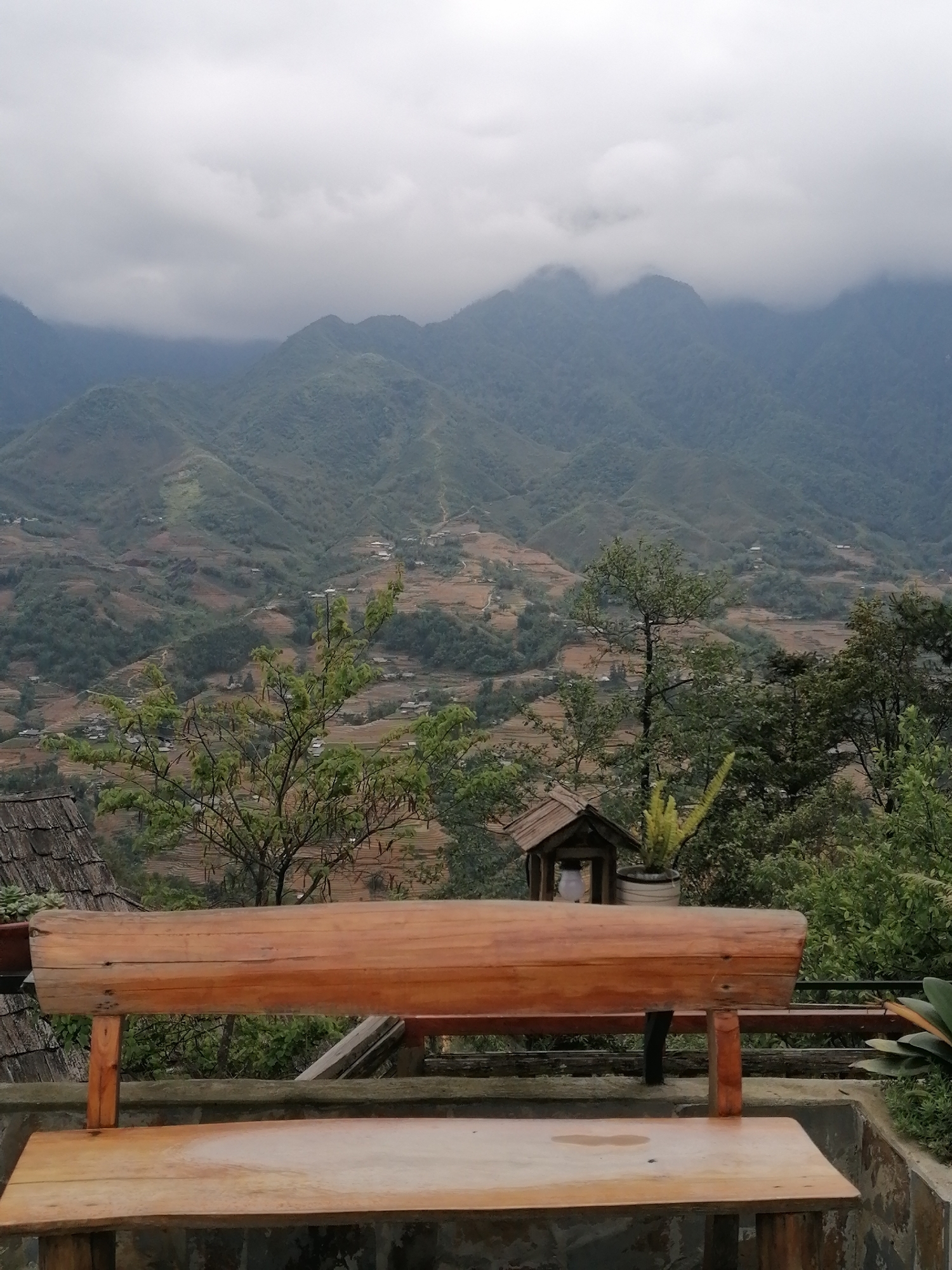 A cloudy vista unfolds from a wooden bench in Sa Pa Ward, Vietnam. Mountain ranges, partially obscured by clouds, form the backdrop. Terraced fields are visible along the mountainsides, dotted with buildings. The foreground contains vegetation, a wooden structure, and a portion of a roof.