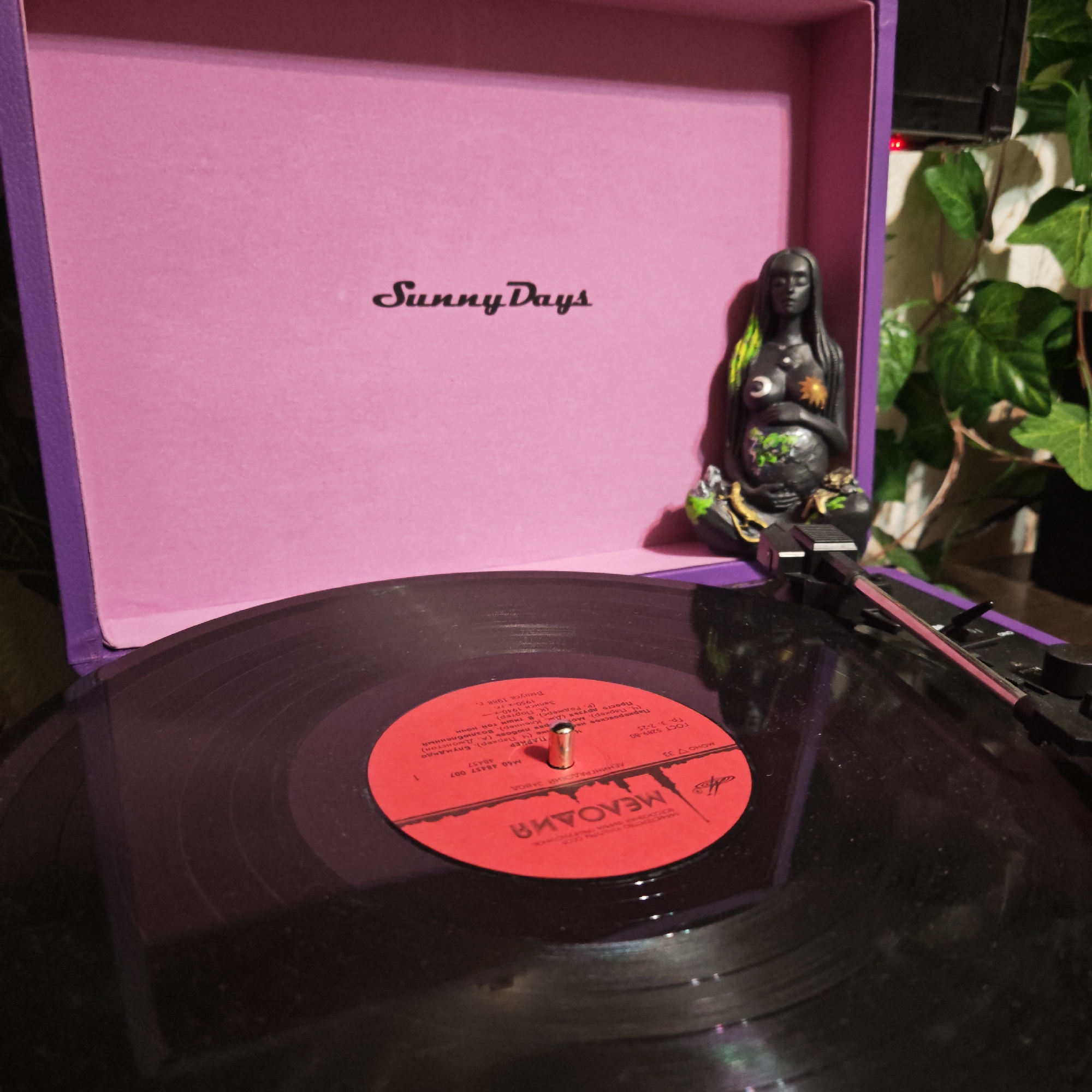 The image presents a close-up, slightly elevated view of a vinyl record playing on a portable record player in Kladovitsy, Russia. The record player's interior lid is pink and features the script text "Sunny Days," while its visible exterior is purple. A black vinyl record with a bright red label is centered on the turntable, with the tonearm and needle positioned, suggesting active playback.
To the right, a striking dark-skinned female figurine is prominently displayed. She has distinctive green and yellow highlights in her long hair, and celestial symbols—a crescent moon on her left breast and a sun on her right—adorn her chest. She cradles a form in her lap, possibly a child or an earth-like creature, also accented with green and yellow. Behind the figurine, lush green, ivy-like foliage is visible, contributing to a natural, somewhat mystic ambiance. The lighting is dim and warm, creating subtle reflections on the shiny surface of the vinyl record.
The red record label contains significant Cyrillic text. It identifies the record company as "МЕЛОДИЯ" (Melodiya), a Soviet-era label, and the manufacturer as "ЛЕНИНГРАДСКИЙ ЗАВОД" (Leningrad Plant). The artist is named "ПАРКЕР" (Parker), with "Ч. Паркер" (C. Parker) specified, strongly indicating jazz musician Charlie Parker. The label notes "Записи 1940-х, 1950-х гг." (Recordings of 1940s, 1950s) and "Выпуск 1988 г." (Release 1988), suggesting a later compilation of earlier jazz works. Other visible details include "ГОСТ 5289-80" (State Standard) and technical numbers like "M60 48437 007" and "МОНО 33".