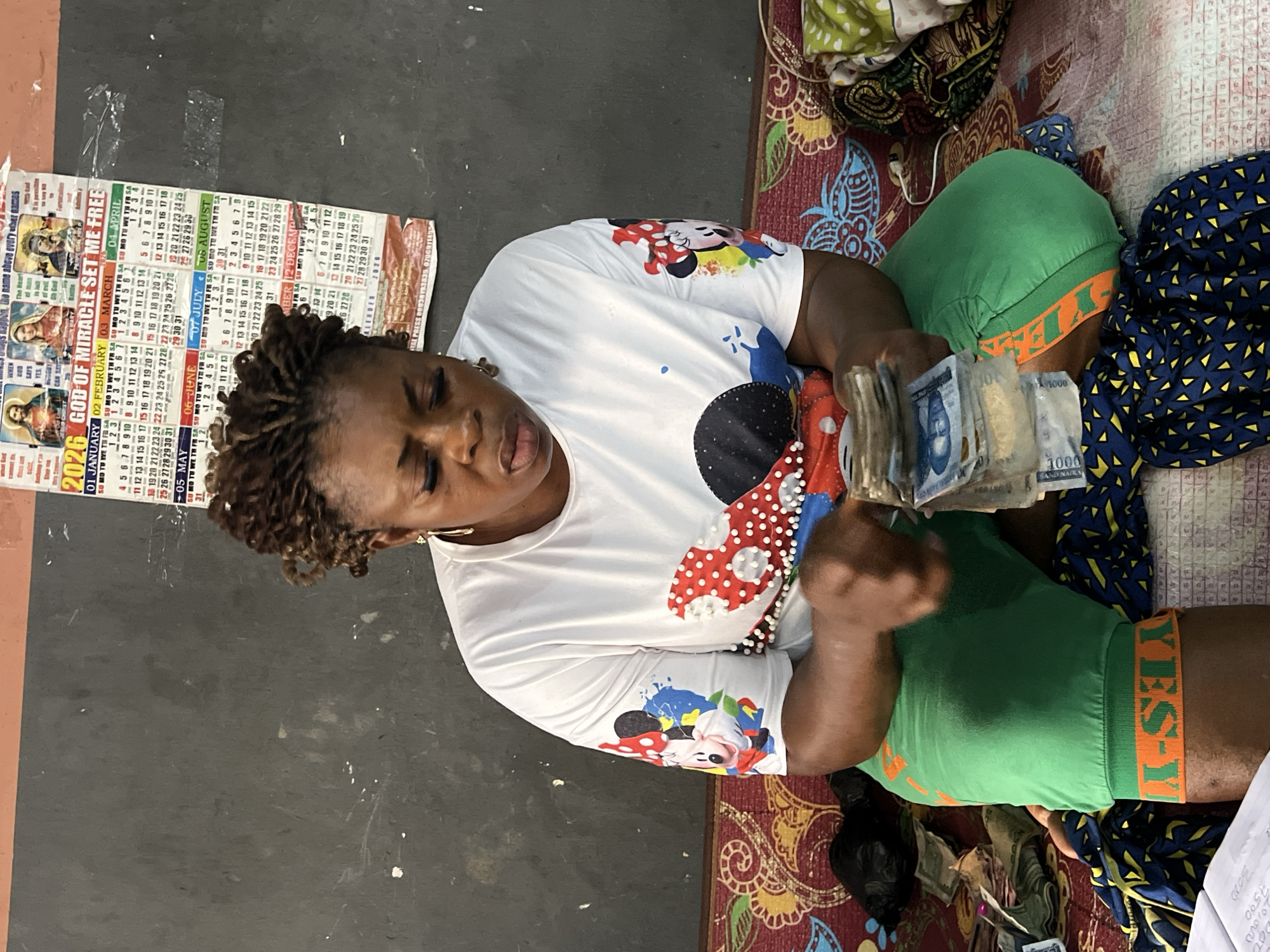 In Nkpor, Nigeria, a woman with braided hair and a white t-shirt adorned with Mickey Mouse characters is seated on a colorful mat. She is wearing green shorts with an orange band that says "YES." The woman is holding a stack of Nigerian currency, appearing to count or organize it. Surrounding her are more piles of cash and some dark fabric. Behind her, a calendar hangs on a dark, textured wall. The calendar is for the year 2026 and has religious imagery and text.