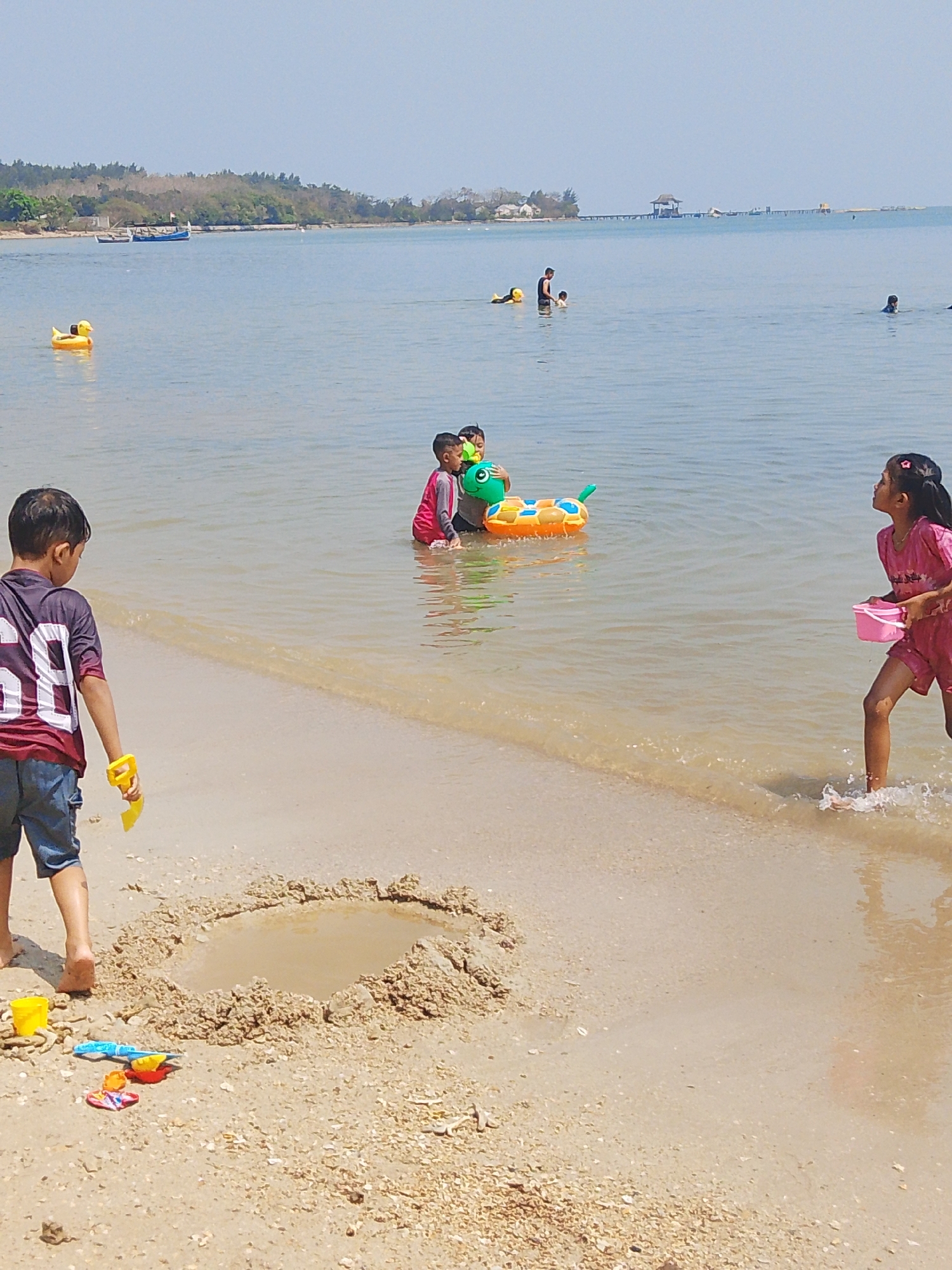 The image captures a sunny day at a beach in Pejagan, Indonesia, where several children are enjoying the water and sand. In the foreground, a young boy in a maroon and grey shirt and blue shorts stands on the sand, holding a yellow toy shovel. Nearby, a small sandcastle has been partially built, with a shallow puddle of water in its center. Several other beach toys, including a yellow bucket, a blue shovel, and colorful molds, are scattered on the sand.
In the shallow water, three children are engaged in play. Two boys, one in a red and pink long-sleeved rash guard, are near an inflatable toy resembling a green caterpillar. Another child is partially visible behind them. Further out in the water, a young girl in a pink dress is running and splashing, holding a pink bucket.
In the background, the calm, clear blue sea extends to the horizon. A few boats are visible in the distance, along with a structure built on stilts over the water. The shoreline is lined with trees and vegetation, indicating a coastal landscape. The overall scene depicts a peaceful, family-friendly environment with children actively participating in beach activities.