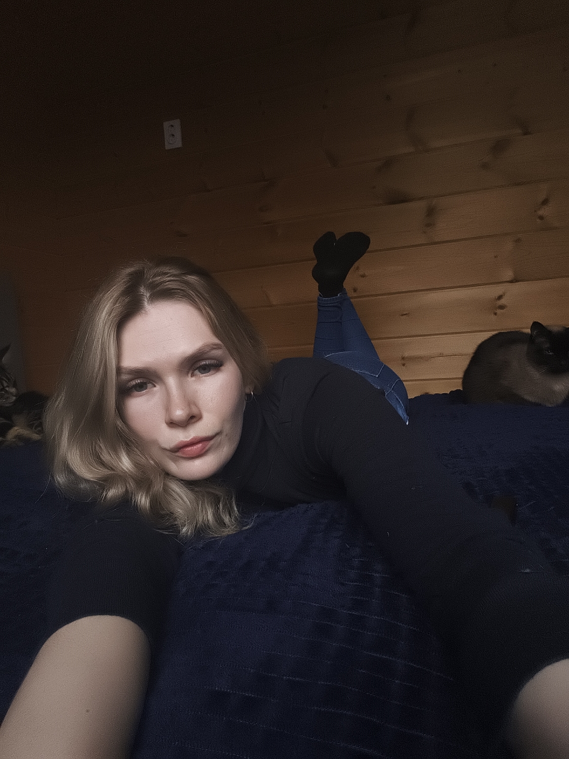 A young woman with blonde hair is captured in a high-angle selfie, lying prone on a dark blue blanket or bedspread. She props herself up on her elbows, looking directly at the camera with a relaxed expression and a subtle pout. She wears a black long-sleeved turtleneck, blue jeans, and black socks, with her bent legs and feet visible in the upper background.
Two cats are also present in the scene. A Siamese-like cat with distinctive light and dark fur is partially visible to the woman's left, near the edge of the frame. Further to the right, a dark-furred cat, possibly black, is resting with its back to the camera.
The setting appears to be an indoor room, characterized by wooden plank walls, which impart a rustic or cabin-like aesthetic. A small white electrical outlet is mounted on the wooden wall in the background. The ambient lighting is somewhat dim, contributing to a cozy and intimate atmosphere. No text is visible in the image. The location context is Русский Вожой, Russia.