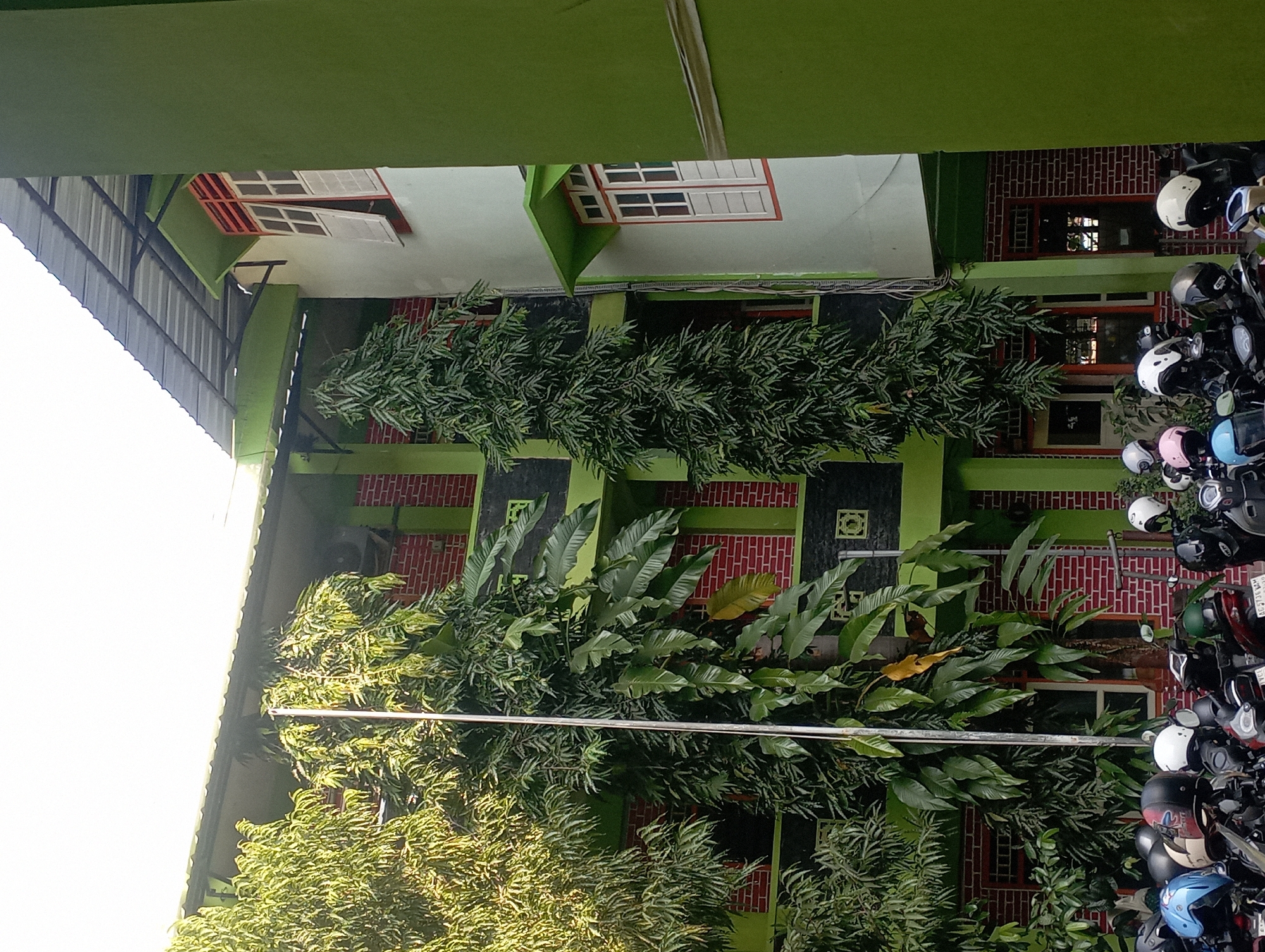 The image shows the exterior of a building in Nganjuk, Indonesia. The building has a light green exterior with dark green accents and red brick walls. There are several windows with white shutters. Lush green trees and foliage surround the building. In the foreground, a row of motorcycles is parked. Some of the motorcycles have helmets on them, including a blue helmet, black helmets, and white helmets. The sky is bright and overcast. The scene appears to be daytime, possibly in the morning or afternoon, given the lighting. There are no people visible in the image.
