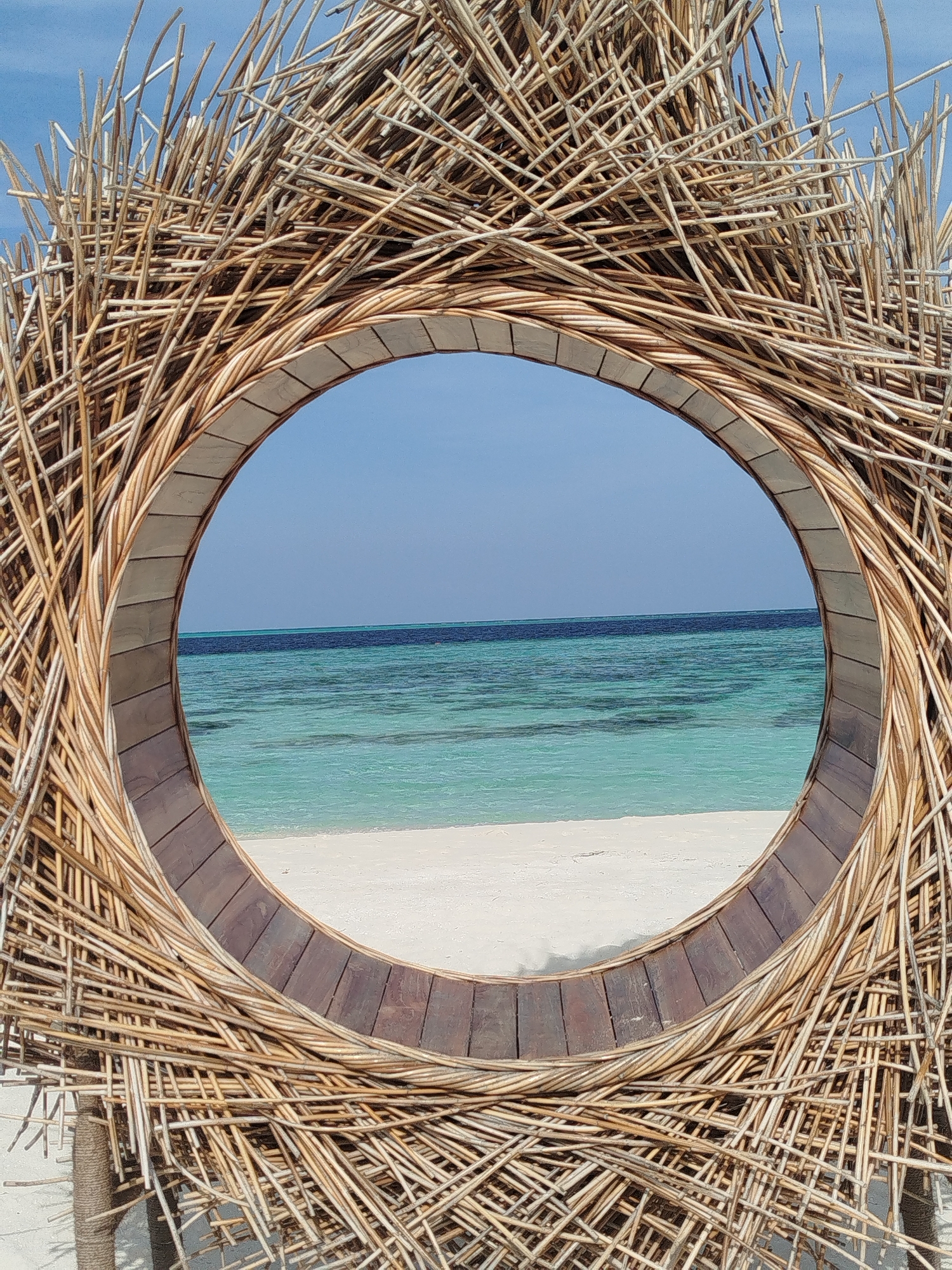 The image captures a circular opening framed by woven natural materials, likely bamboo or reeds, creating a rustic, nest-like structure. Through this opening, a serene view of the ocean and beach in Hinnavaru, Maldives, is visible. The water transitions from a light turquoise near the shore to a deeper blue further out. A strip of white sand beach is present between the water and the foreground structure. The sky above is a clear, light blue. No people are present in the scene, and the focus is on the natural landscape and the art installation.