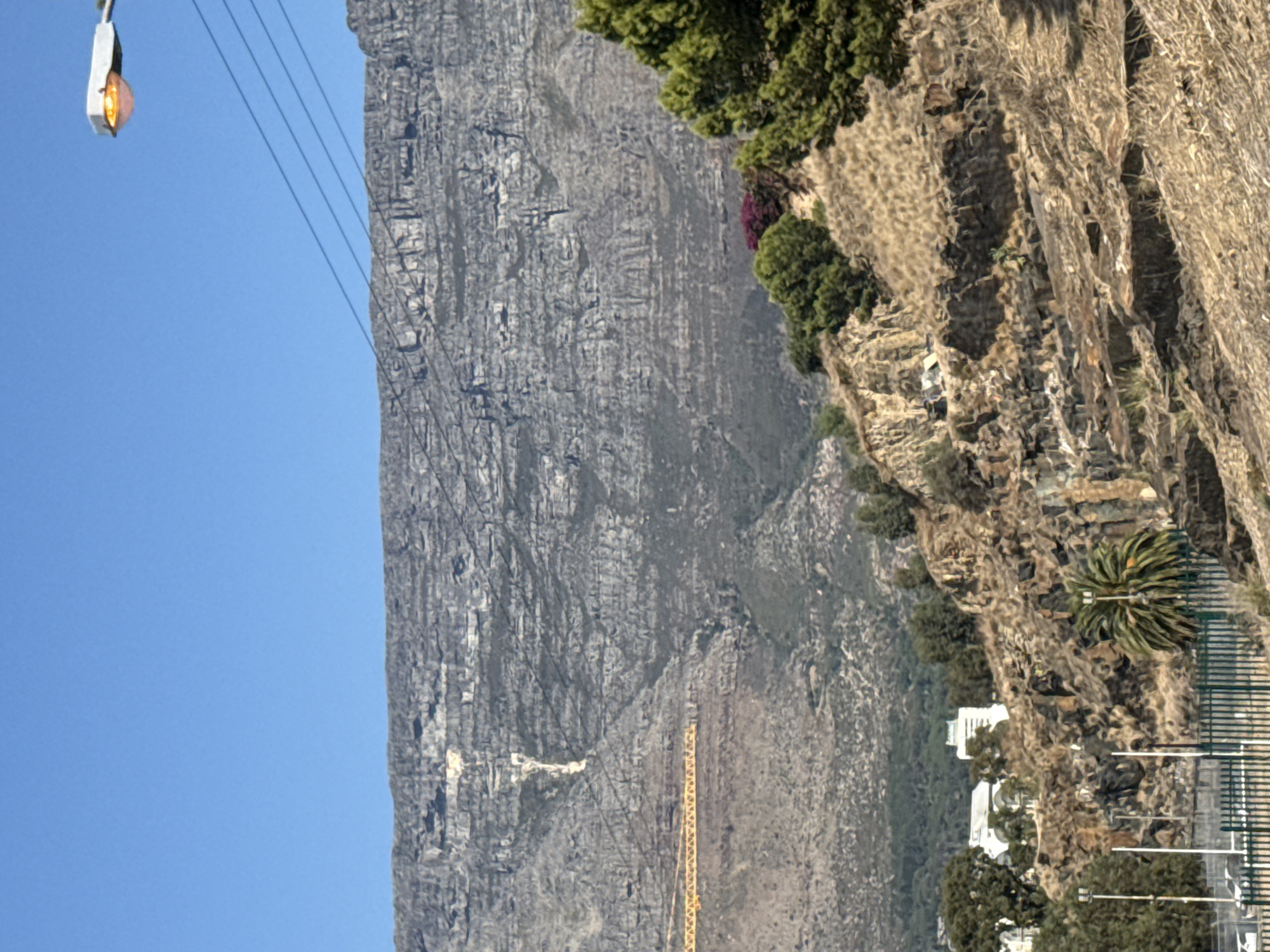 The image shows Table Mountain in Cape Town, South Africa, with a clear blue sky in the background. A cable car wire stretches diagonally across the image, and a light hangs from it at the top left corner. The mountain's rocky face is visible, and vegetation grows on the slope to the right. Buildings and a fence are visible at the base of the slope. A construction crane is visible on the face of the mountain.