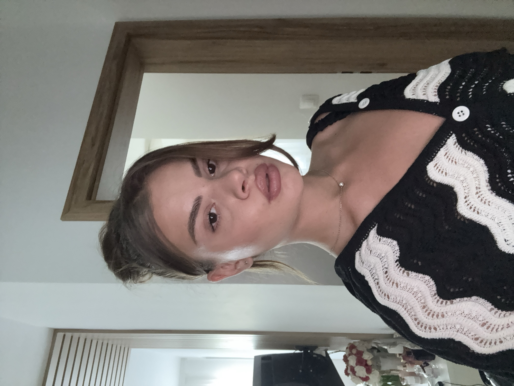 The image displays a young woman posing for a photo, looking directly at the camera with a slight pout and her head tilted. Her dark brown hair is pulled back, with a few strands framing her face. She is wearing a black knitted top or cardigan with distinct white wavy patterns and white buttons. A thin, silver-colored necklace with a small, sparkling pendant is visible around her neck, and she has a small stud earring in her ear. Her lips appear to be accentuated with a mauve-pink color, and her eyebrows are well-defined. The background is an indoor setting with a light-colored wall and a wooden door frame partially visible. In the lower right portion of the image, some indistinct objects can be seen, including what looks like a dark electronic device and a small arrangement of white and red artificial flowers. The lighting is soft and even. No visible text is present.