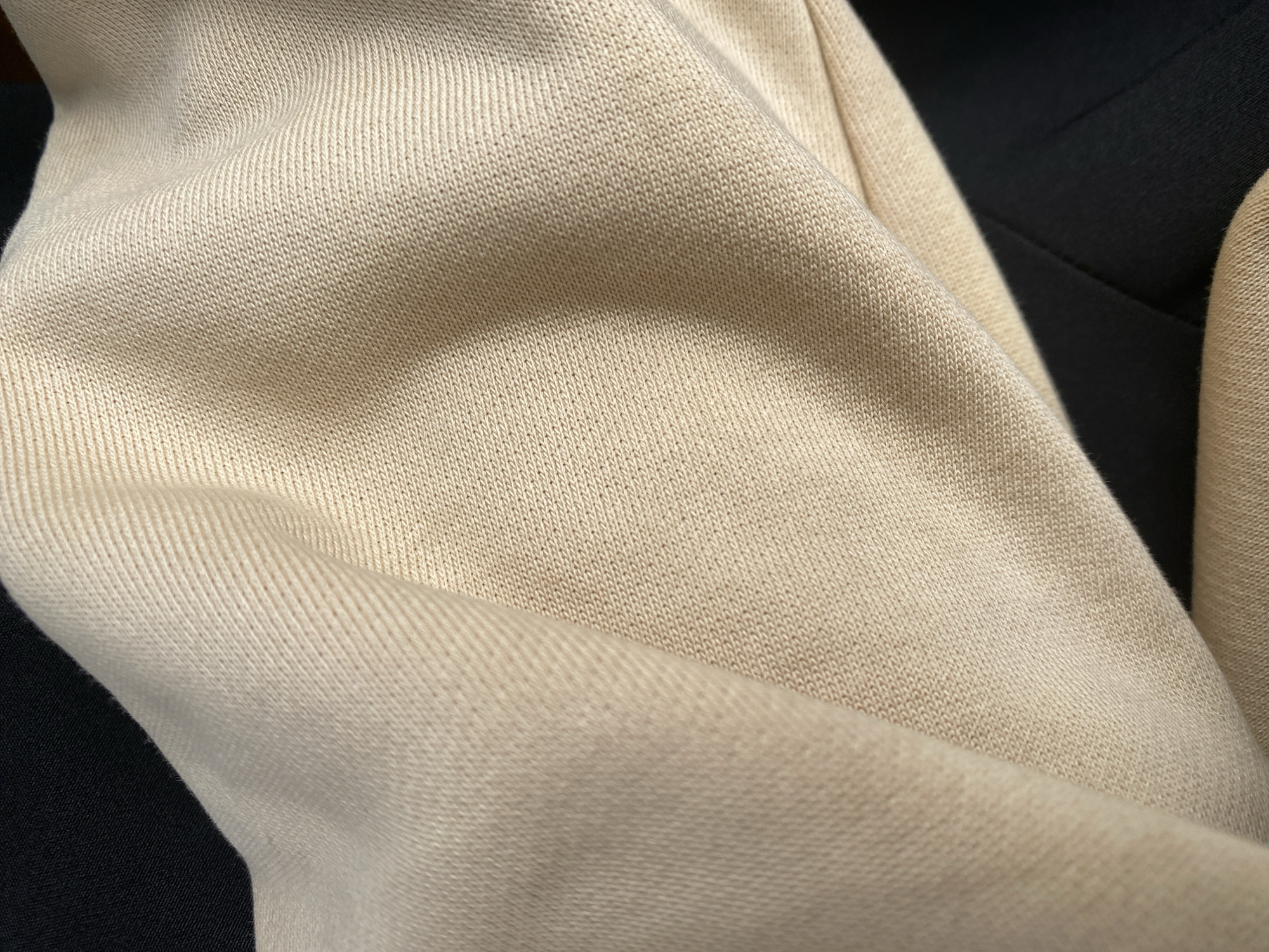 This is a close-up, overhead shot of a cream-colored, knitted fabric, possibly a garment. The fabric is draped, creating folds and shadows that highlight its texture. The knitting appears to be a fine gauge with a ribbed pattern. To the right and background, a darker fabric, likely black, is visible, suggesting the cream fabric is part of a larger item or placed on top of the black material. The lighting is soft and diffused, indicating an indoor setting or an overcast day. There are no people, specific location cues, or text visible in the image. The focus is entirely on the texture and drape of the fabric.