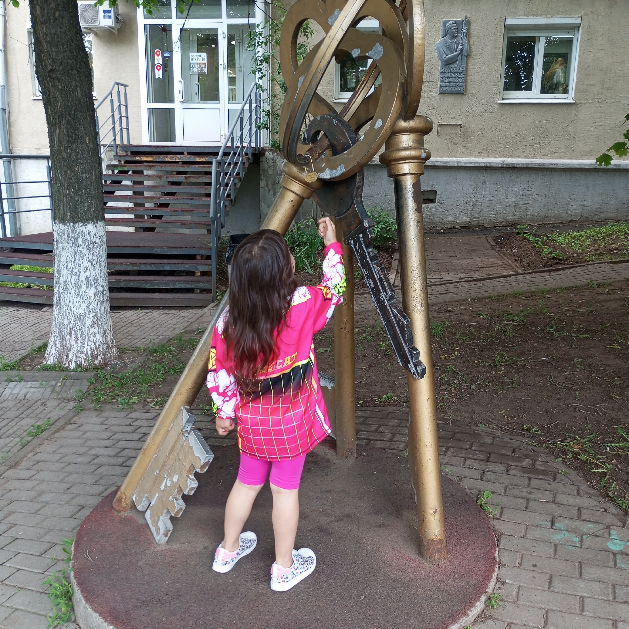 A young girl with long brown hair, wearing a pink outfit and patterned sneakers, stands in front of a large golden sculpture of intertwined keys. The sculpture is situated on a circular base on a brick-paved area, with a tree to the left and a building with stairs and a doorway to the background. The building appears to be an office or administrative building, as suggested by the sign on the door that reads "Реклама Открыто" (Advertising Open) and business hours. A plaque with a portrait of a man is mounted on the wall of the building. The overall scene suggests an outdoor urban setting. This sculpture is located near the building of the Udmurt State University in Izhevsk, Russia, on the corner of Universitetskaya Street and Studentcheskaya Street.