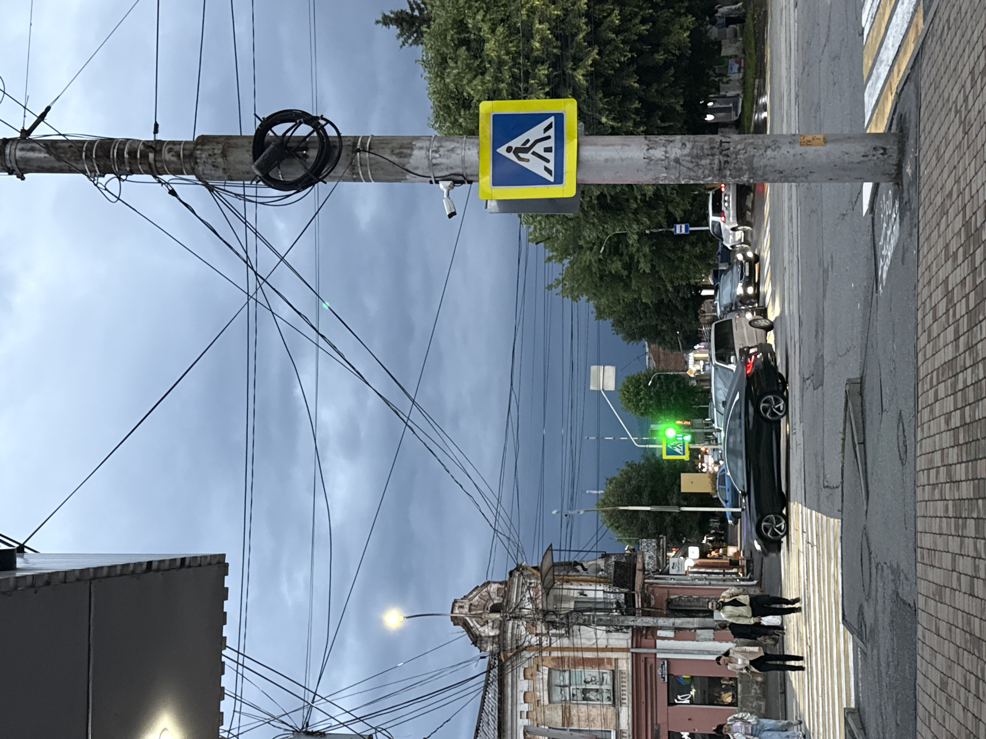 A street scene in Assinovskaya, Russia, shows a cloudy, overcast day, likely late afternoon or early evening. The image is oriented vertically. A prominent utility pole, laden with numerous electrical wires, dominates the left side of the frame. Attached to this pole is a traffic sign for a pedestrian crossing, a blue square with a white pedestrian symbol, bordered by a yellow square. A security camera is also mounted on the pole.
In the background, a road with a pedestrian crossing marked by yellow and white stripes is visible. Several cars are driving on the road, with headlights on. A traffic light is showing green. On the right side of the road, a building with a weathered facade and ornate architectural details stands. Across the street, more trees and buildings can be seen.
Several people are visible on the pedestrian crossing in the foreground, appearing to be walking. The overall mood is somber, likely due to the overcast weather. The image lacks any easily discernible text, except for what might be graffiti on the utility pole and building facades, which is too indistinct to read.