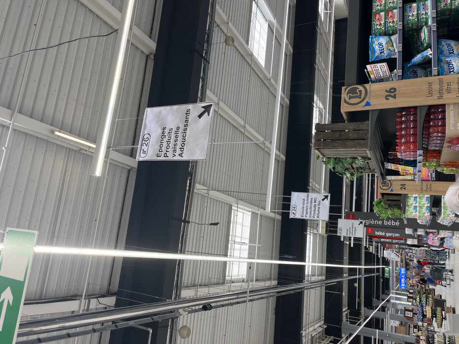 The image shows the interior of a supermarket in Saint-Brice-Courcelles, France. The location is marked by the coordinates: latitude 49.283, longitude 3.999. The image focuses on a sign hanging from the ceiling, with the number "26" printed on it. The sign reads, "Éponges Produits vaisselle Adoucissants" (Sponges Dishwashing Products Softeners) and points to the right with a black arrow. The sign is located above aisle number 26.
There are numerous other signs hanging from the ceiling, indicating other product locations, such as aisle number 24 which features "Essuie-tout Papters toilette Incontinence Produits PVC Allume feu" (Paper towels Toilet paper Incontinence PVC products Fire starter).
There are also signs with the letter "L" in a circle, potentially the logo for the supermarket chain. The aisle in the image is filled with various cleaning products, mostly bottles of red liquid, stacked on shelves, as well as a few cleaning supplies and a sign for "Lessive Nettoyant vitres Linettes" (Laundry detergent Glass cleaner Linens). The environment appears to be brightly lit and modern, with a metal ceiling grid, metal beams, and skylights. The camera angle is pointing slightly upward towards the ceiling. It is difficult to determine if there are any people in the image, due to the low image resolution.
