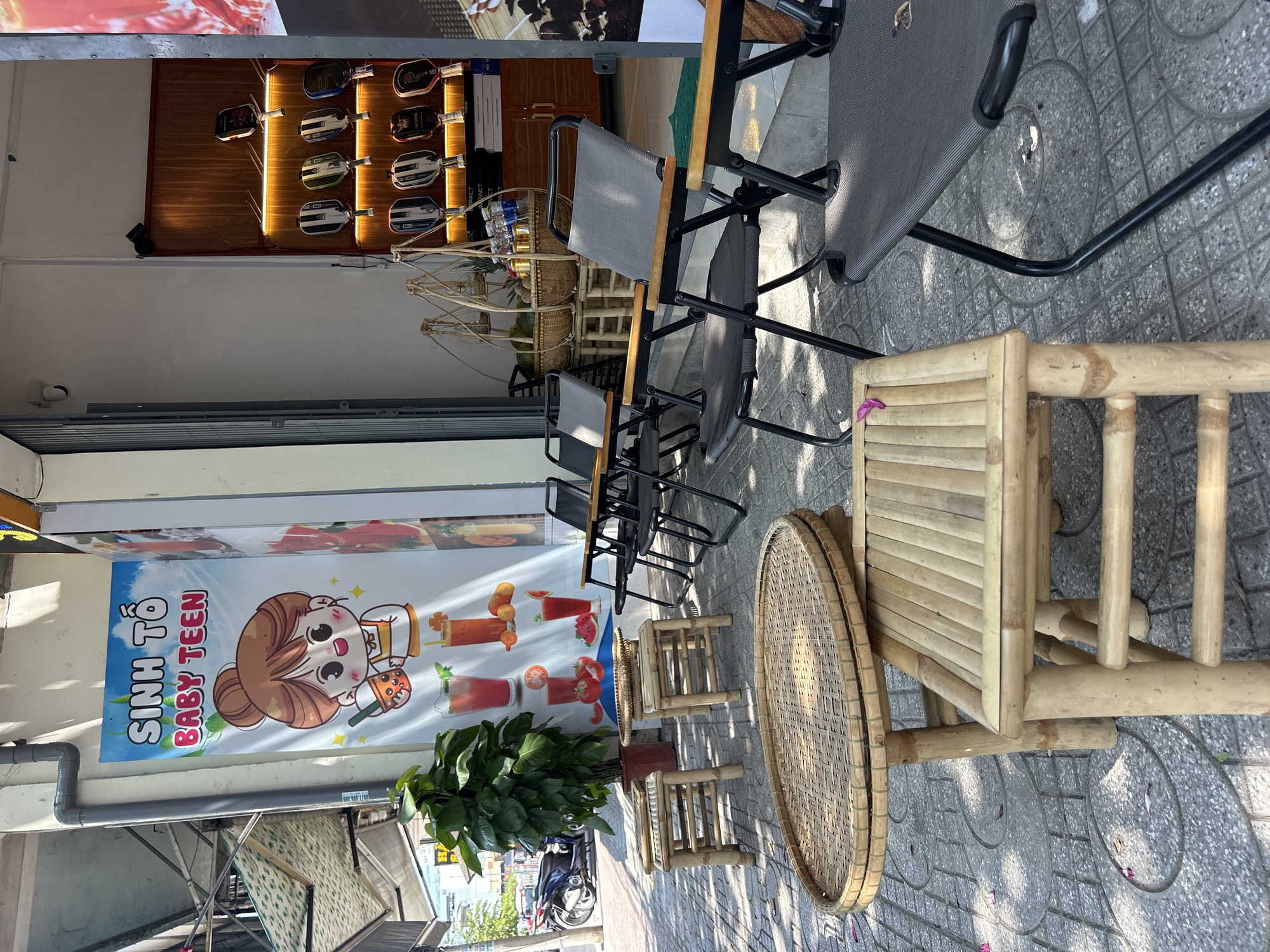 The image depicts an outdoor scene in Nha Trang, Vietnam. A shop selling drinks, advertising "Sinh To Baby Teen" (Smoothie Baby Teen) is visible. The sidewalk displays an arrangement of outdoor seating. Several metal-framed, gray camping chairs and small bamboo tables and stools are scattered on the gray-paved sidewalk. A large potted plant sits in front of the drink advertisement. A display of racing equipment on the wall above the chairs, and motorbikes are parked along the curb in the street.